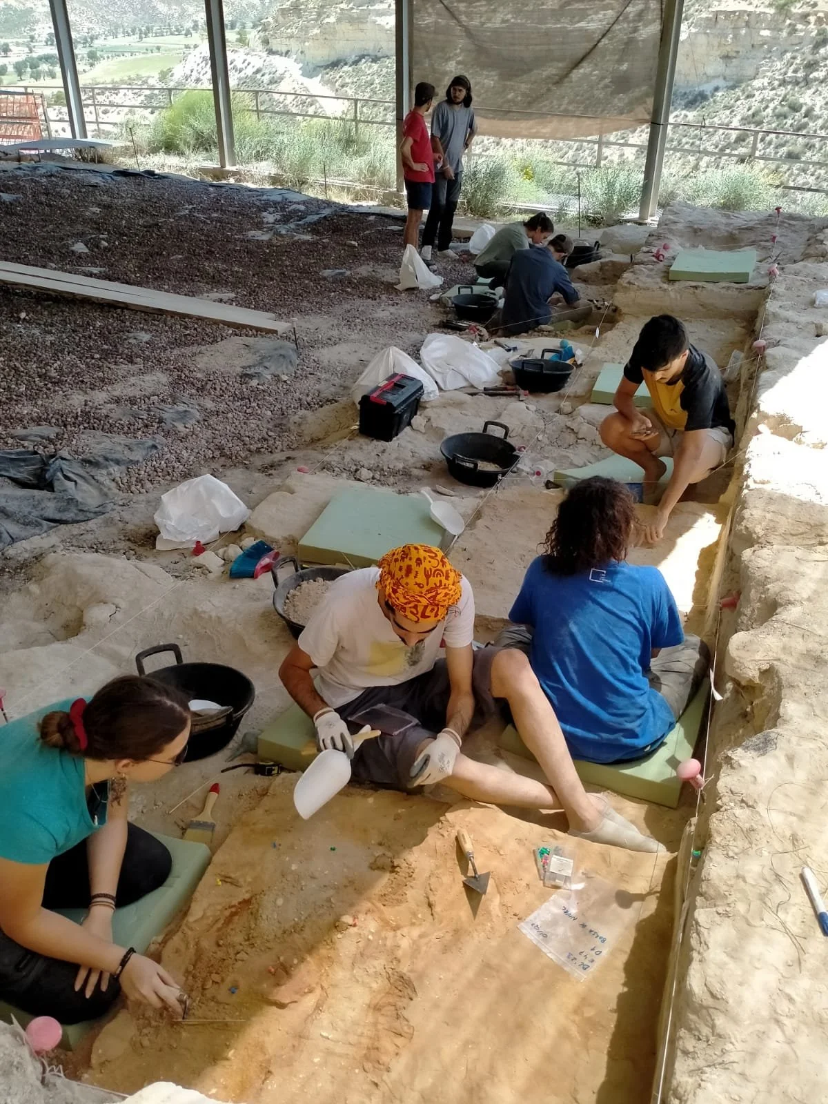 Archaeologists working at an excavation site with tools, sitting and standing around trenches with exposed ancient artifacts.