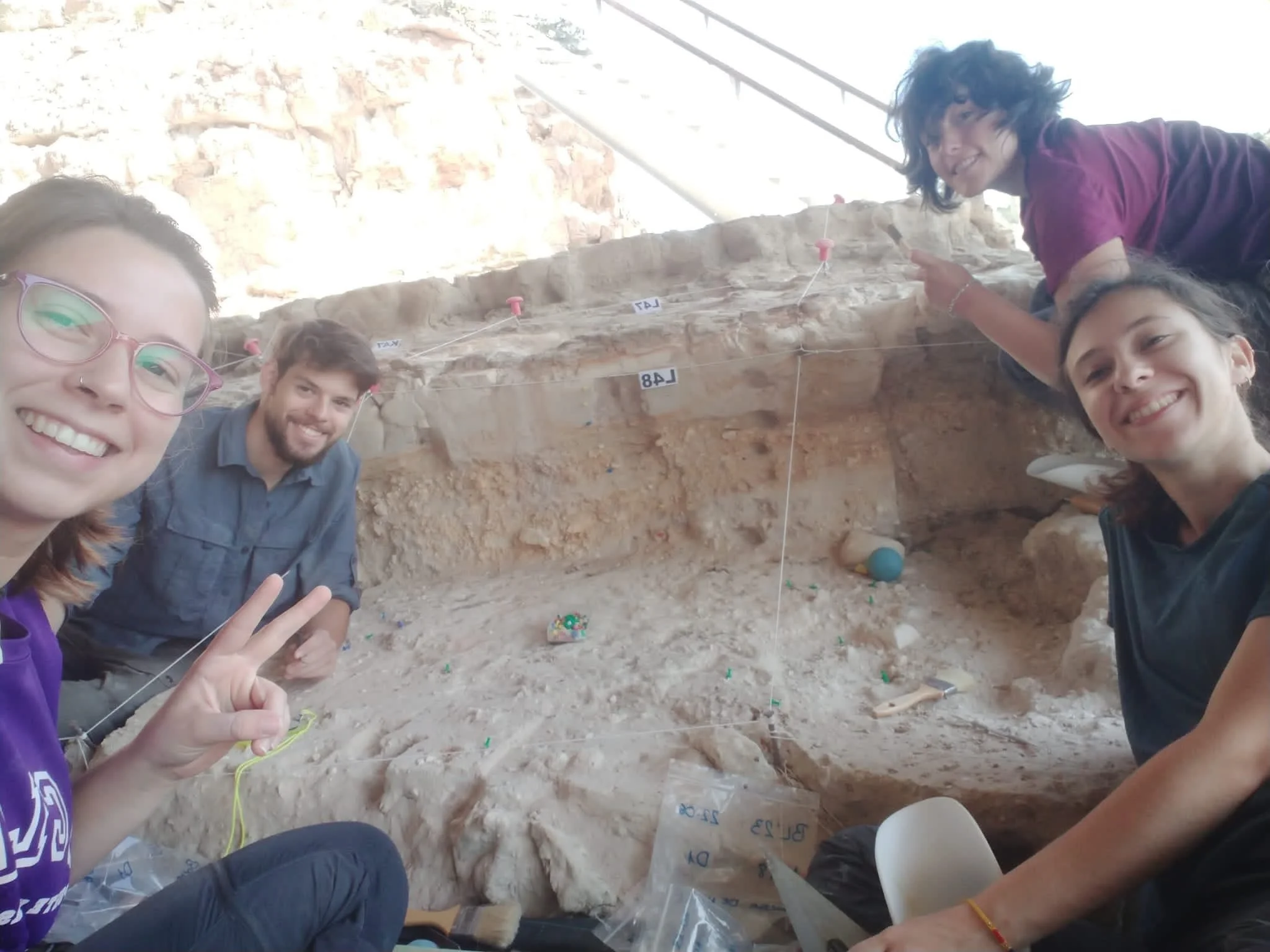 Group of five people smiling and working on an archaeological excavation at a site with exposed layers of earth and rocks.