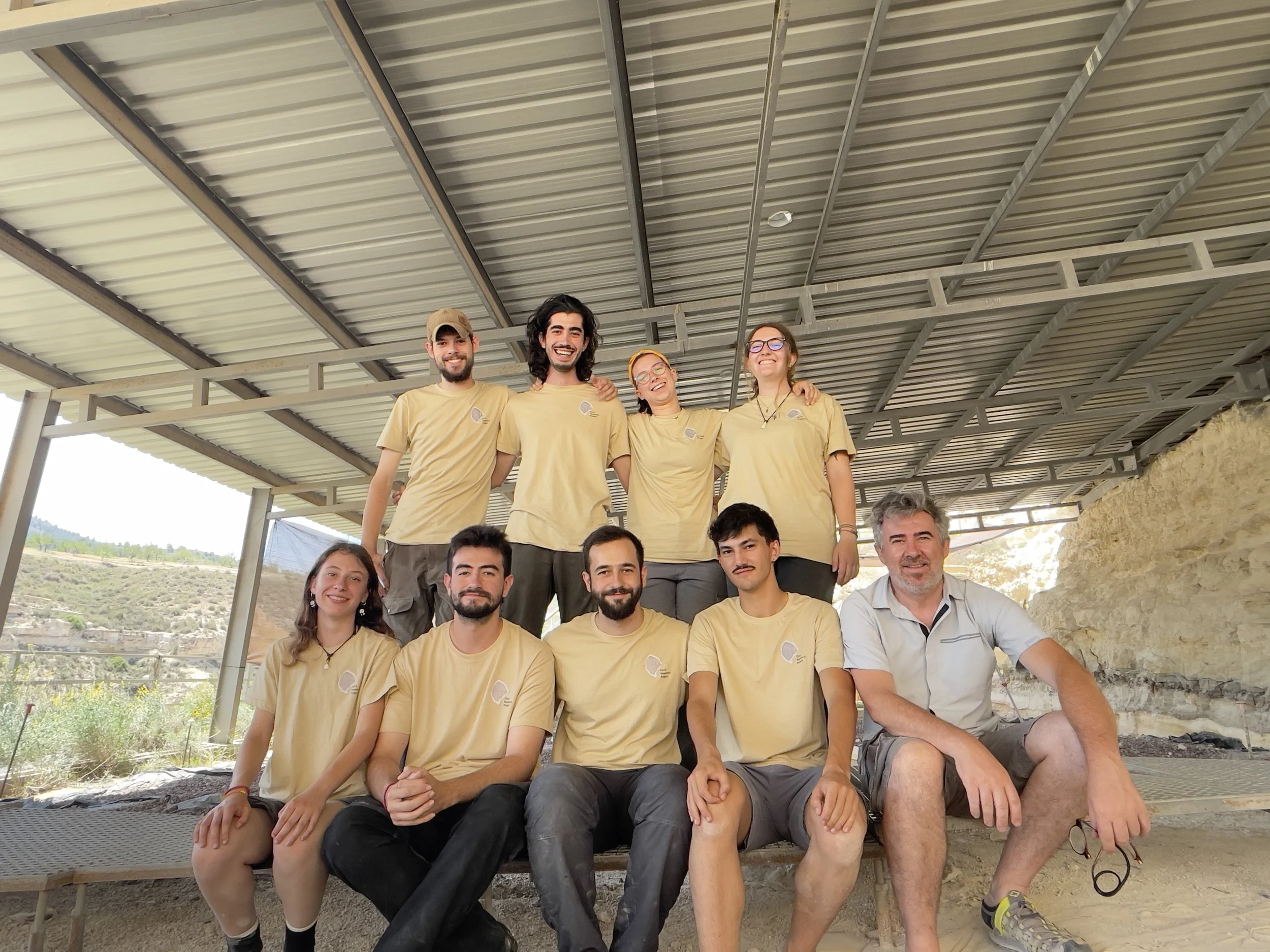 A group of ten people, wearing matching beige shirts, posing under a metal roof structure outdoors. Some are standing, some are sitting, and they are smiling.