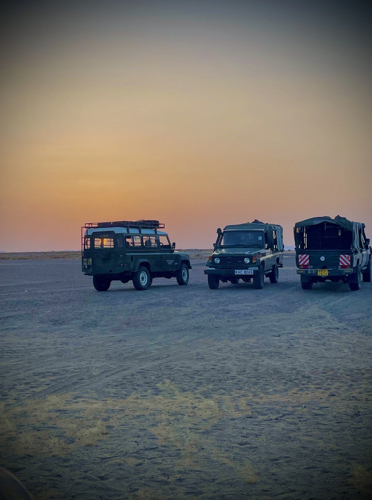 Three off-road vehicles parked on sandy terrain during a sunset or sunrise with a colorful sky.