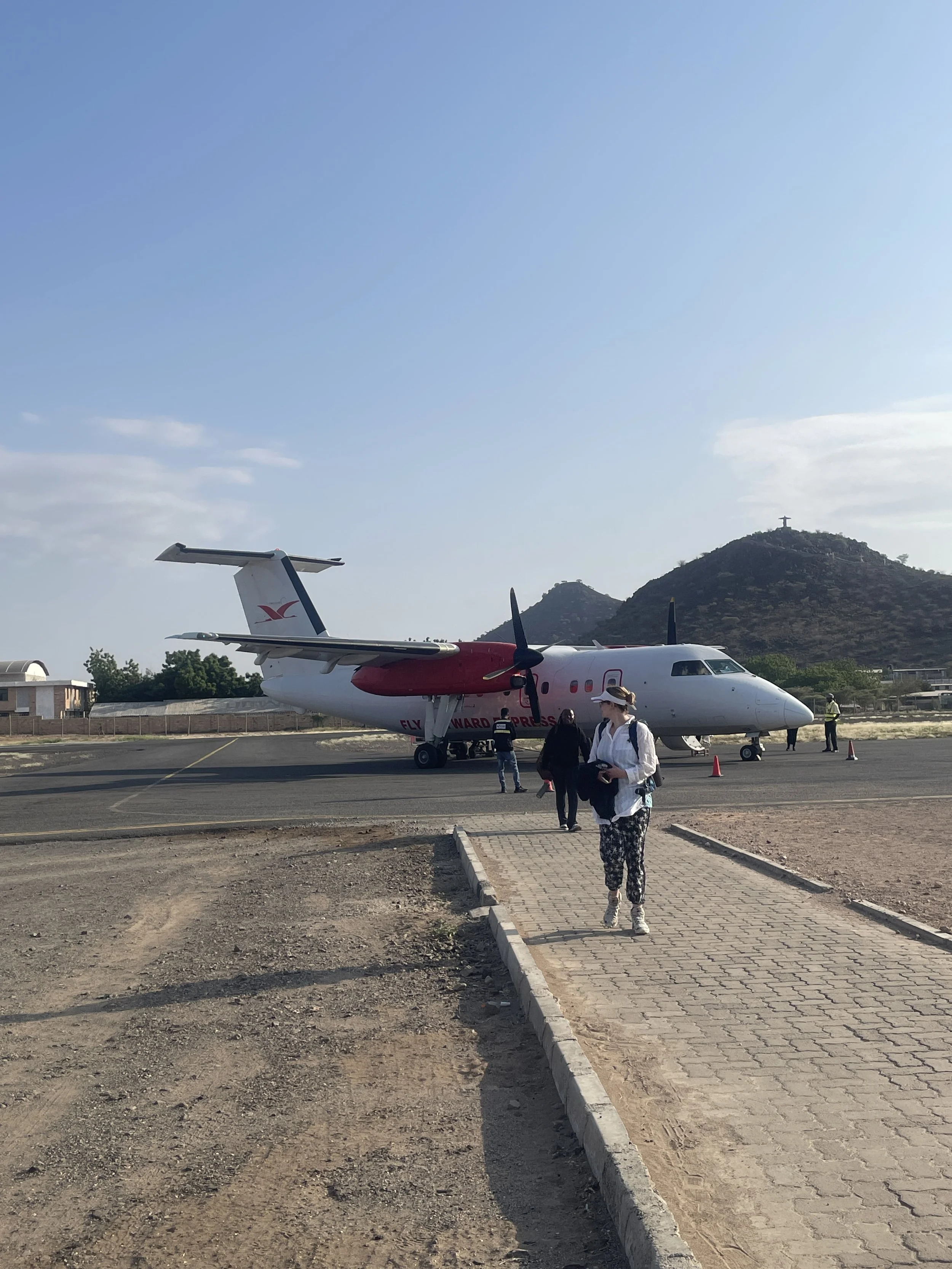 People walking towards a small airplane on a tarmac with hills and a lighthouse in the background.