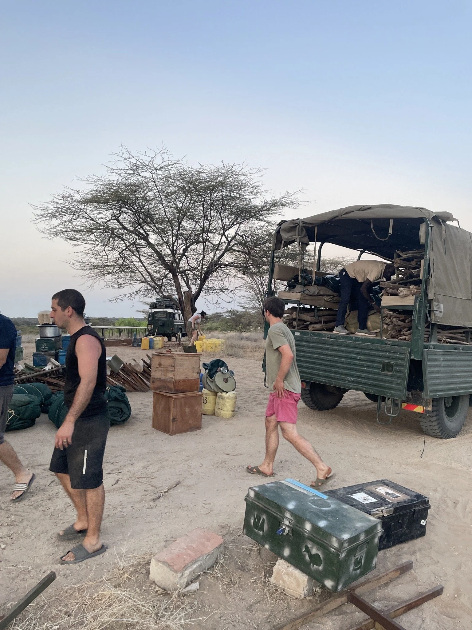 Group of people setting up equipment near a large truck in a dry, flat landscape with a leafless tree under a clear sky.