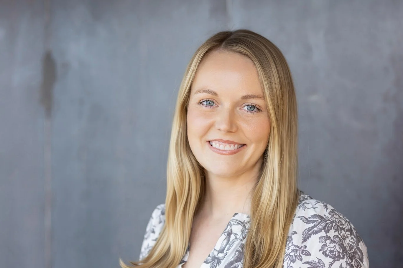 A smiling woman with long blonde hair and blue eyes, wearing a white blouse with a floral pattern, against a gray background.