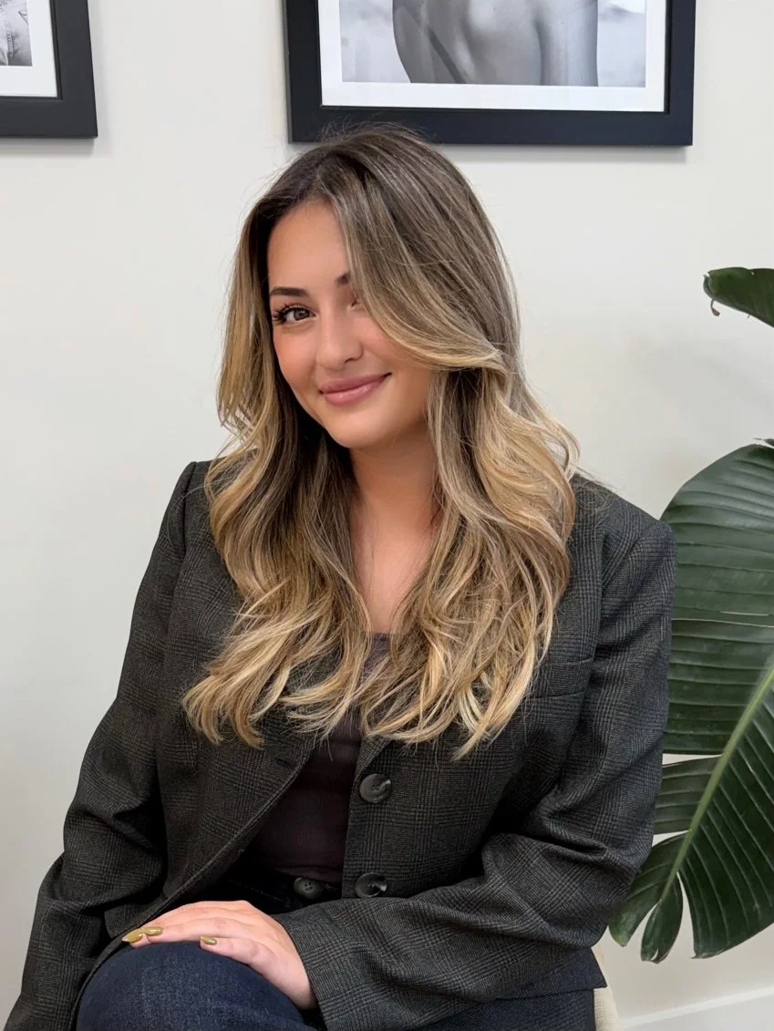 A woman with wavy, shoulder-length hair sitting on a chair, smiling at the camera, in a professional setting with framed black-and-white artwork on the wall behind her and a large green plant nearby.