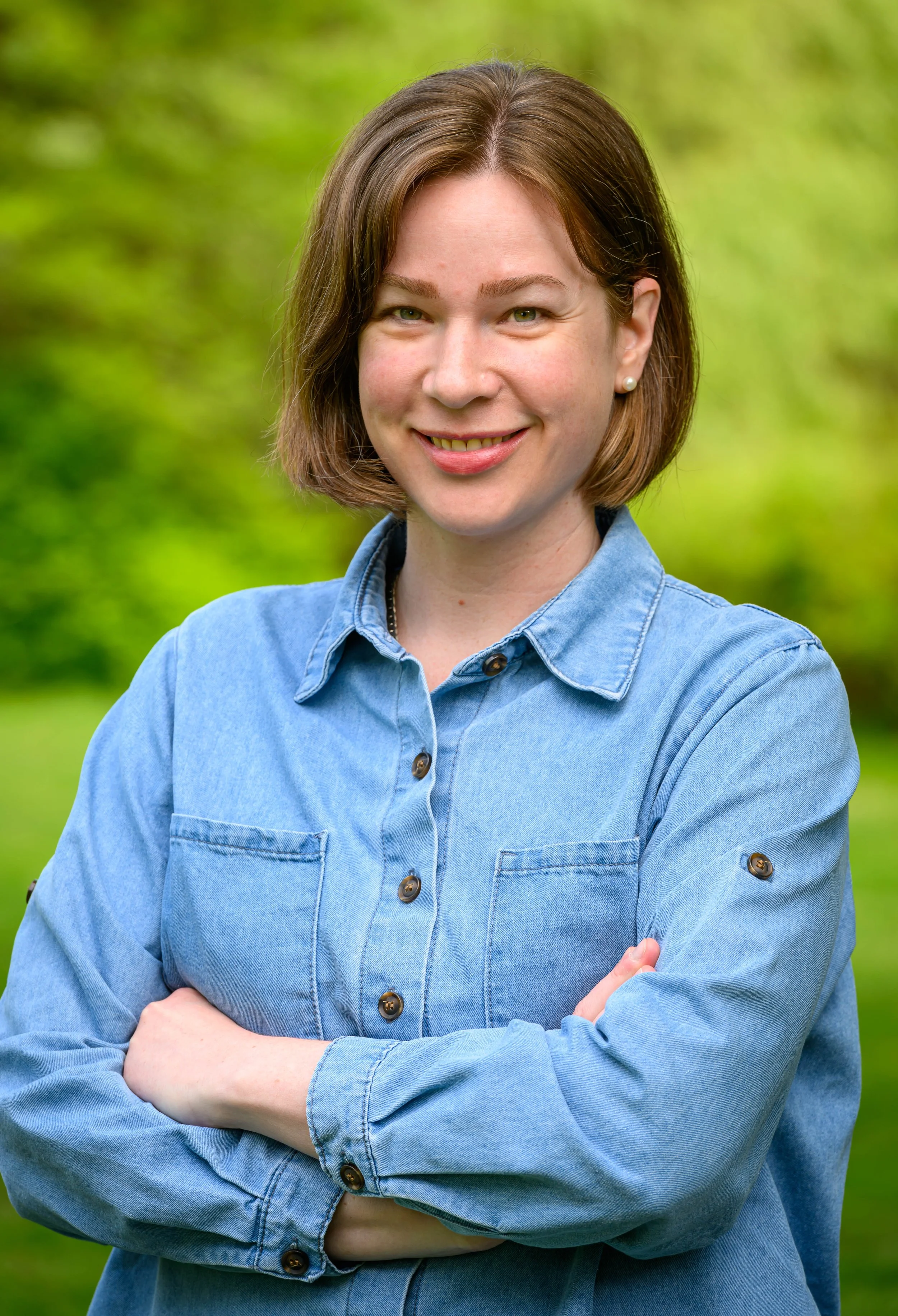 A woman with shoulder-length brown hair, wearing pearl earrings and a blue denim shirt, standing outdoors with her arms crossed and smiling at the camera against a blurred green background.