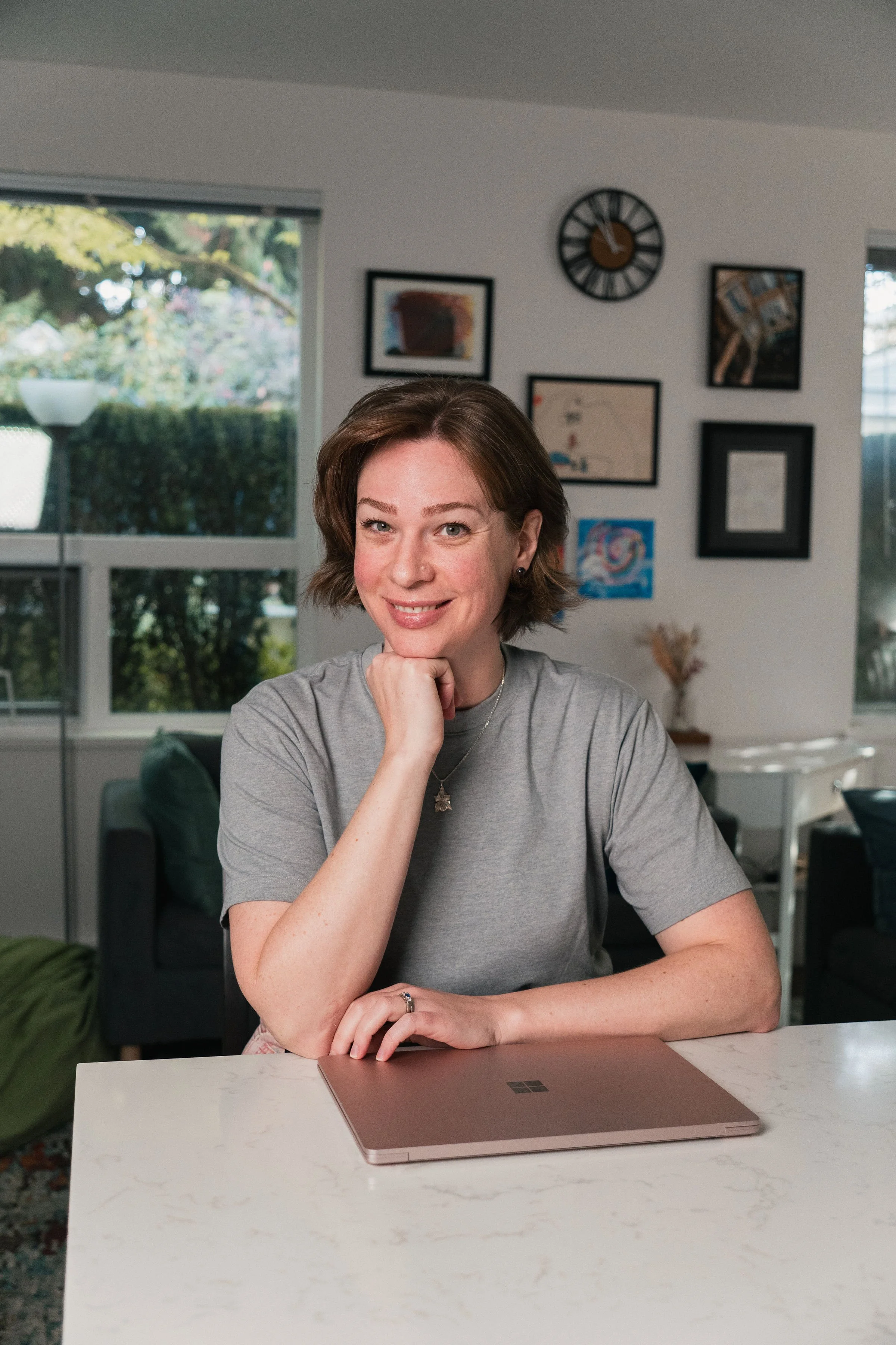 A woman with short brown hair smiling, resting her chin on her hand, sitting at a table with a closed rose gold laptop in front of her in a modern home with wall art and a large window showing greenery outside.