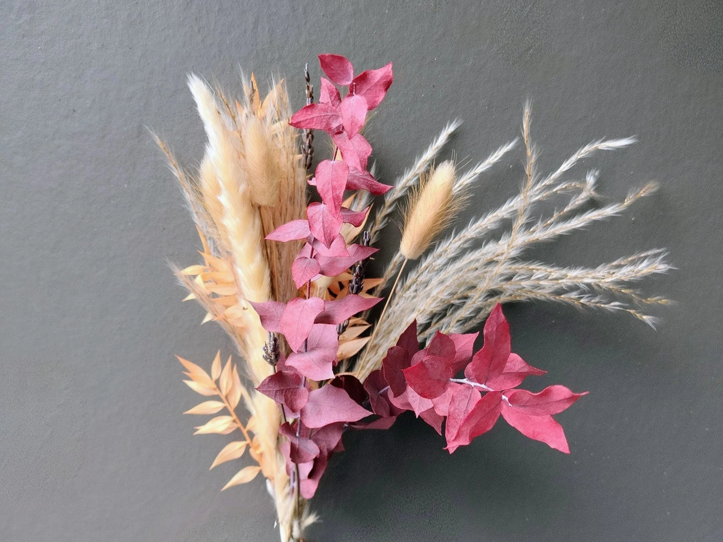 A bouquet of dried flowers and grasses, including pink leaves, beige pampas grass, and other dried foliage, arranged on a plain gray background.