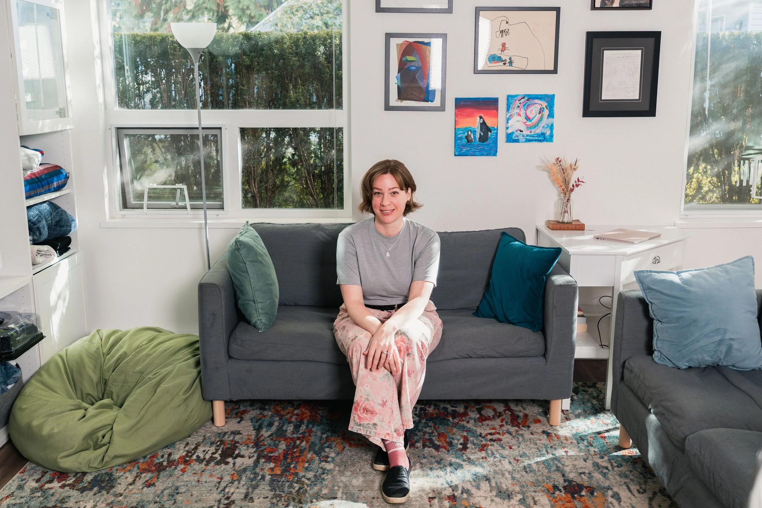 A woman sitting on a dark gray sofa in a living room, smiling, with colorful pillows; large windows with greenery outside; wall with framed artwork; a white side table with a dried flower arrangement; patterned rug on the floor; a green bean bag on the floor.