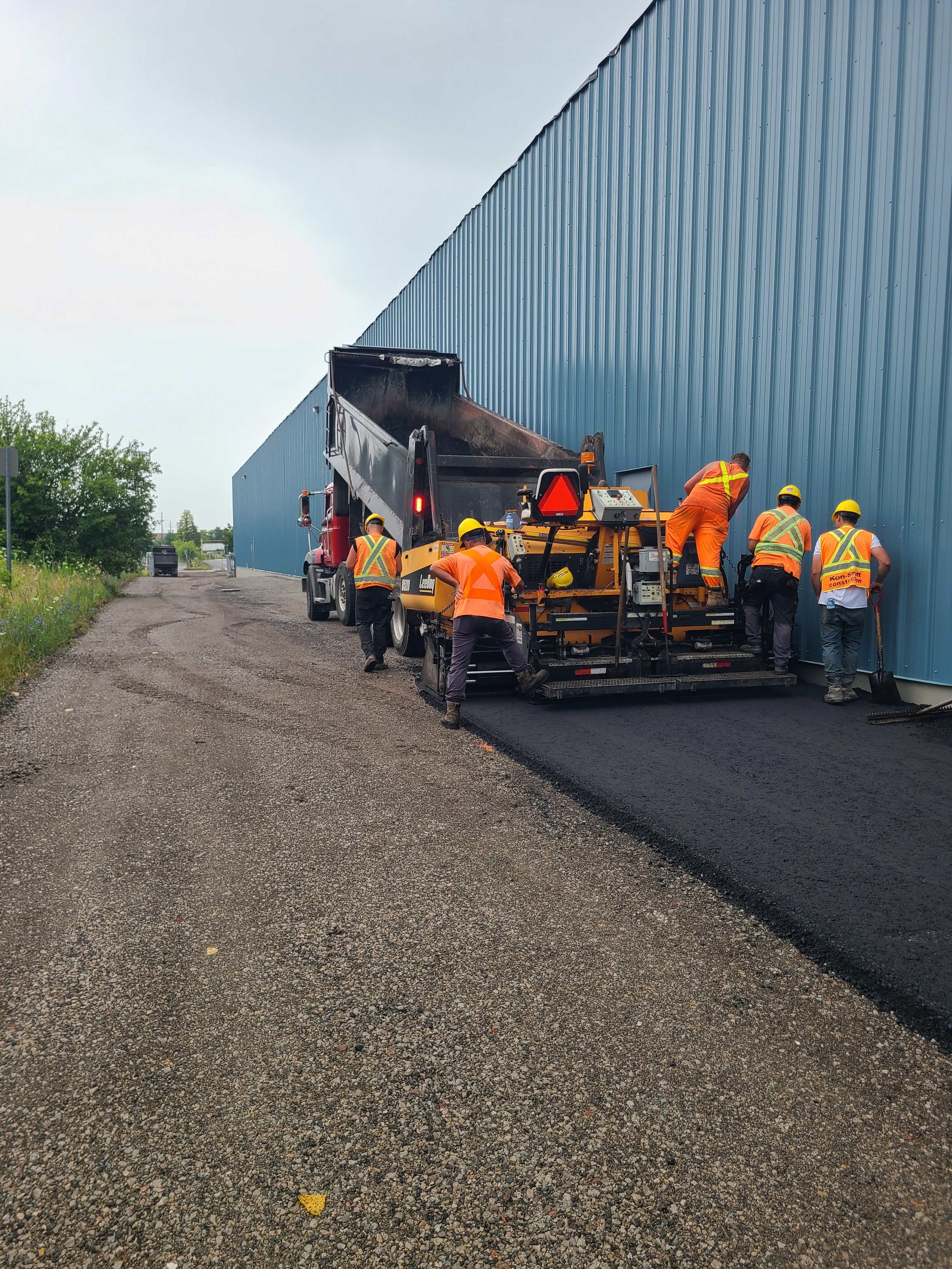 Workers in orange safety vests and yellow helmets laying asphalt on a construction road next to a large blue warehouse building.