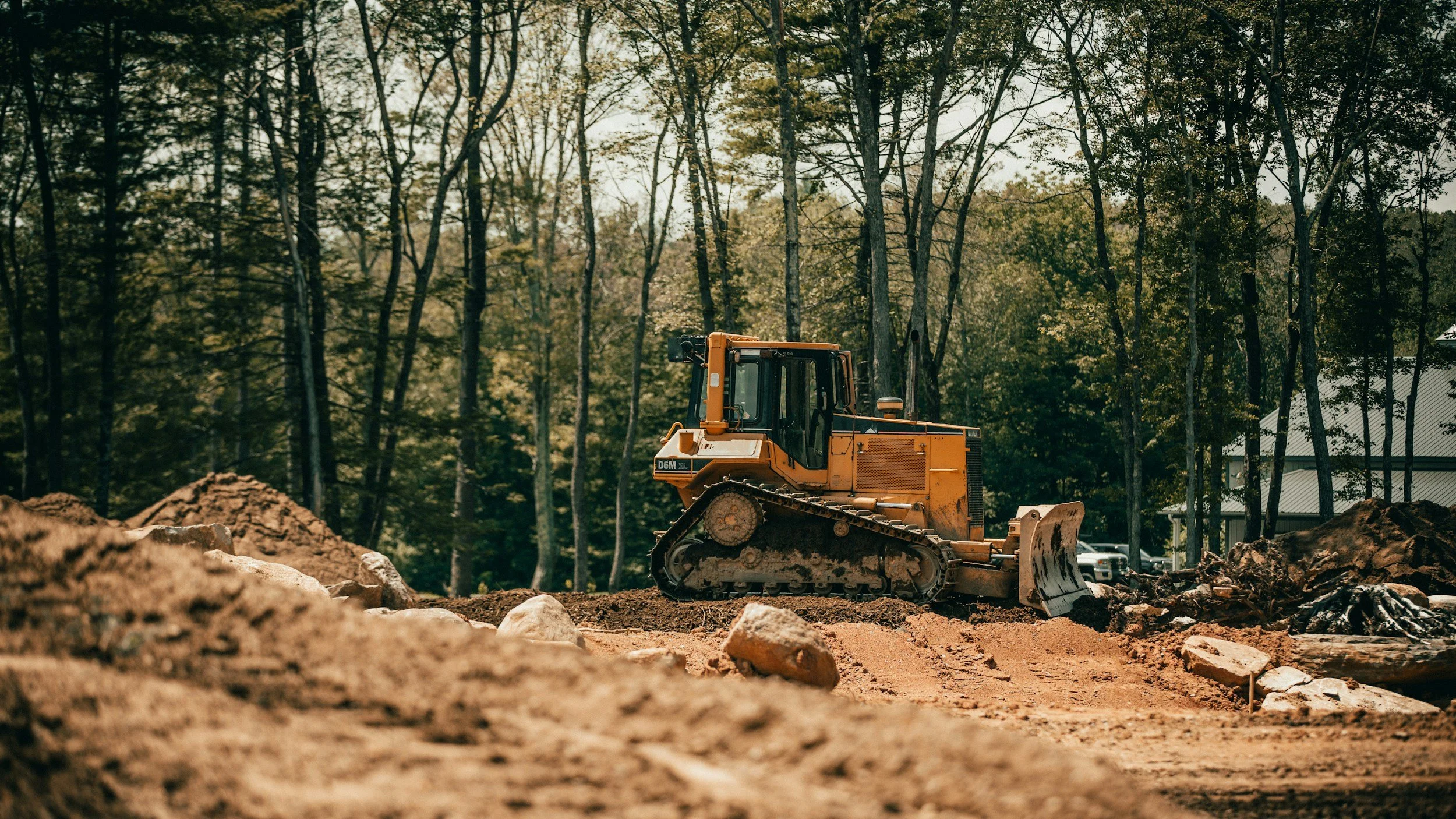 A bulldozer working on a dirt construction site surrounded by trees with a building in the background.