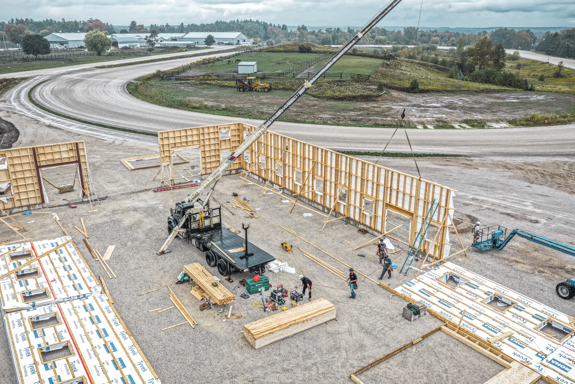 Construction site with workers building wooden frame walls, construction materials, tools, and a crane lifting a wall section. A gravel road curves in the background with rolling fields and trees.