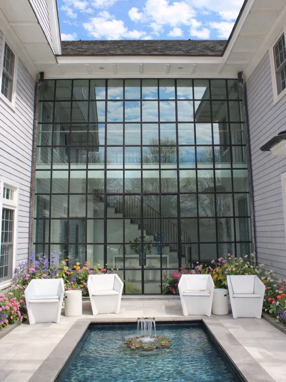 Indoor courtyard with a small rectangular fountain surrounded by chairs, colorful flowers, and a large glass window reflecting the sky and trees.