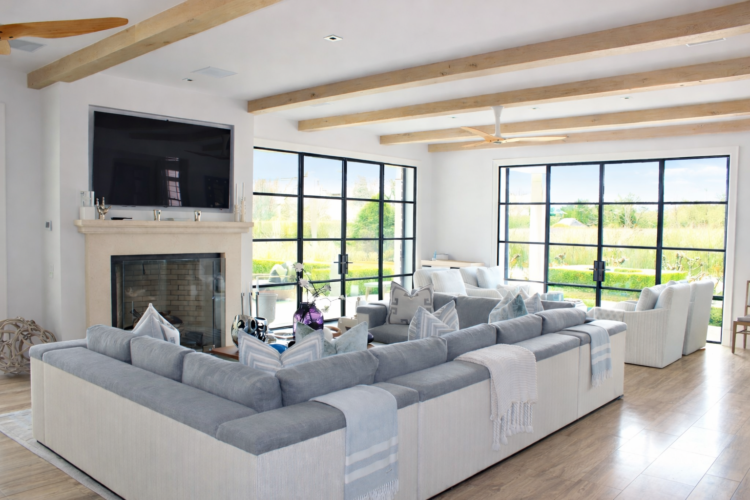 Bright living room with large windows, gray and white sectional sofa, white armchairs, wooden ceiling beams, fireplace with TV above, coffee table with decor, and a ceiling fan.