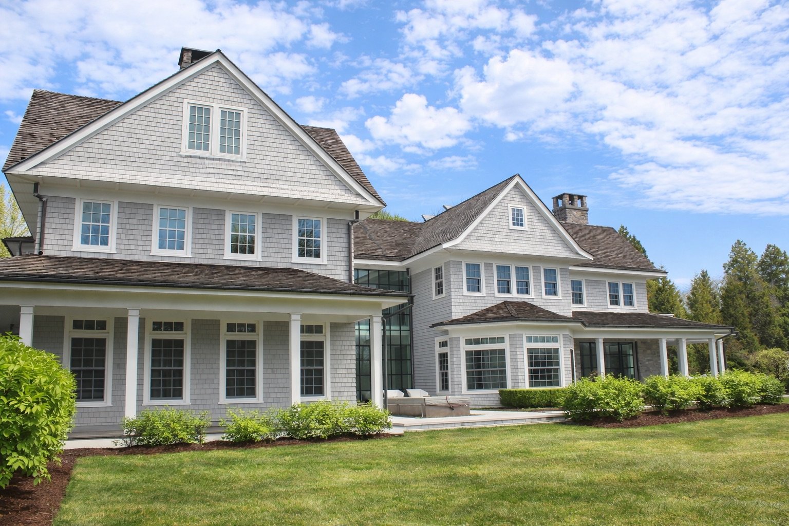 A large white house with multiple gabled roofs, numerous windows, and a wraparound porch, set against a backdrop of blue sky with clouds and surrounded by green lawn and trees.
