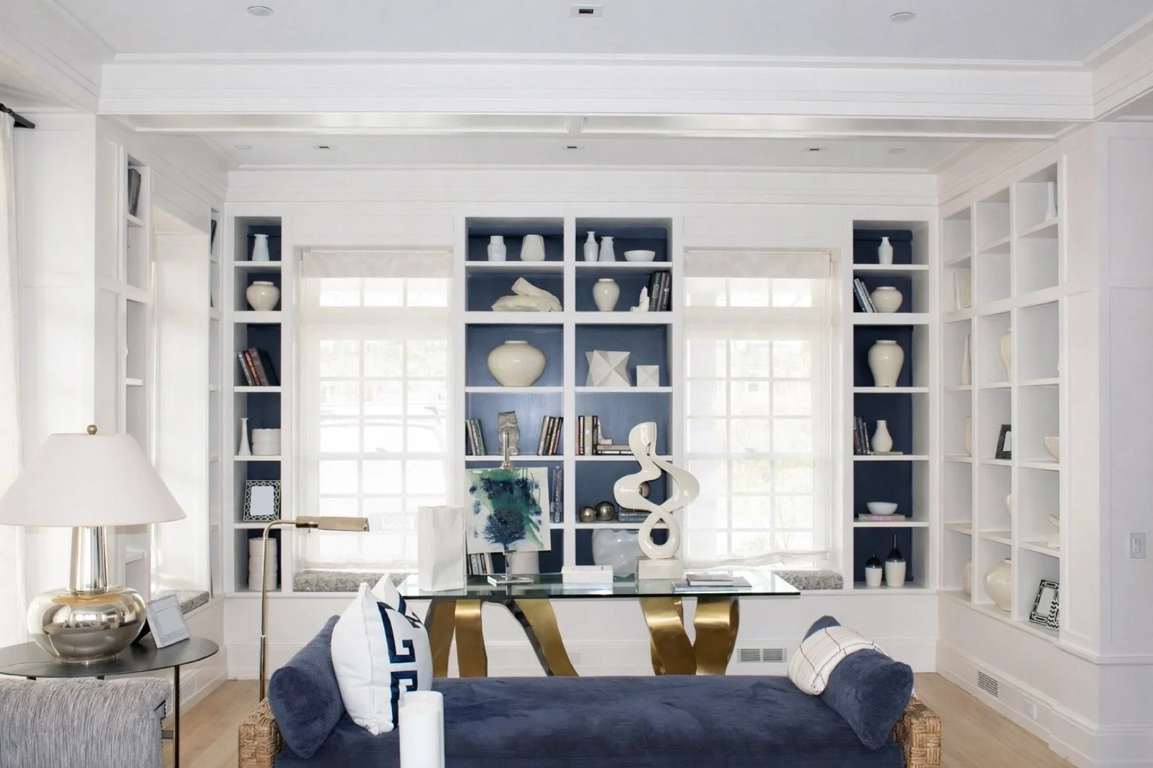 Living room with built-in white bookshelves, large windows, and a glass table, decorated with white and dark blue accents.