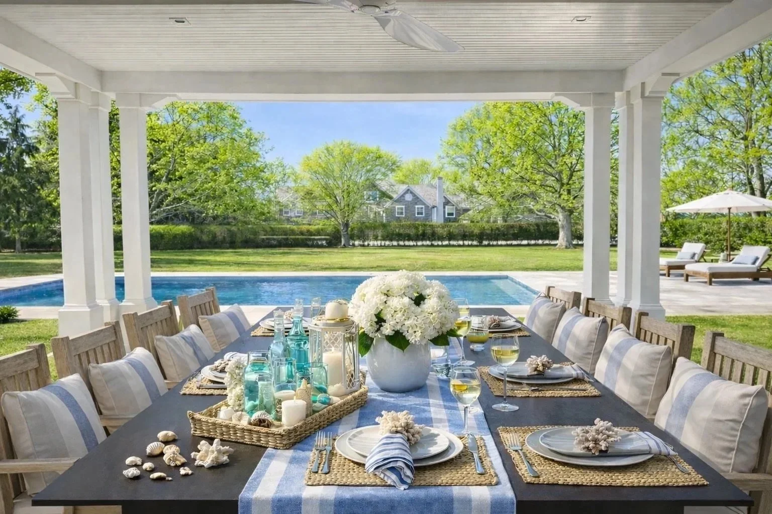 Outdoor dining table set with seashells, candles, and glassware, overlooking a backyard pool and lush green trees on a sunny day.
