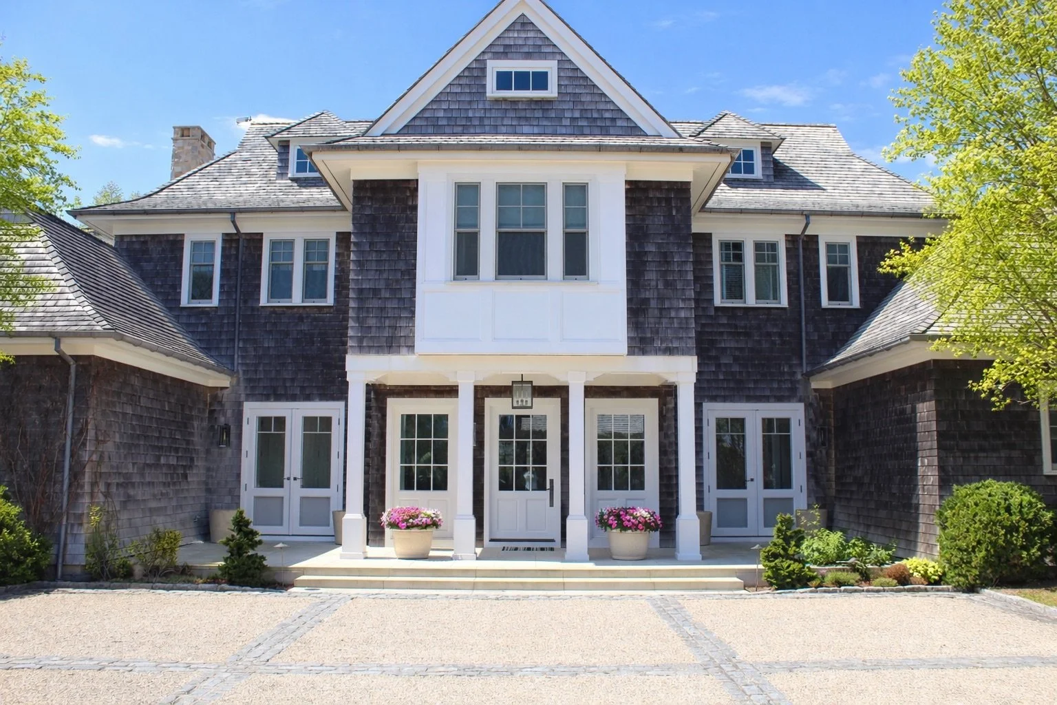 Front view of a large house with dark wooden siding, multiple windows, and white trim. There is a porch with steps, potted flowers, and a walkway leading to the entrance. Trees and a blue sky are in the background.