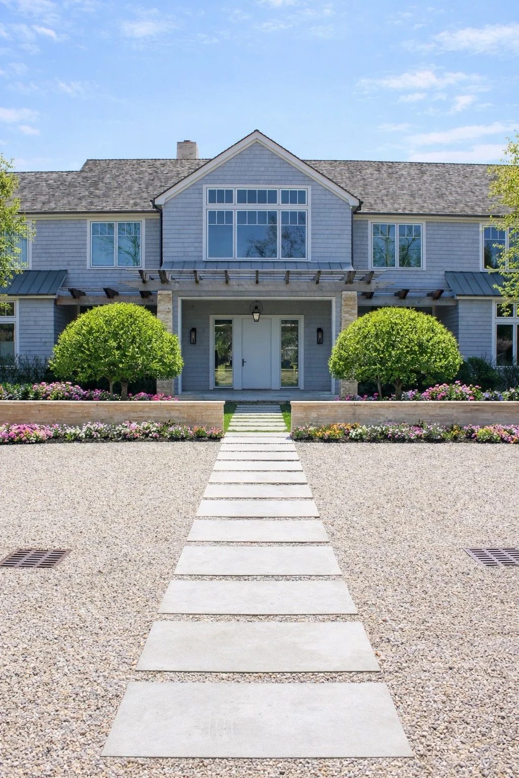 Front view of a modern house with a paved walkway leading to the front door, surrounded by landscaped bushes and flower beds under a clear blue sky.