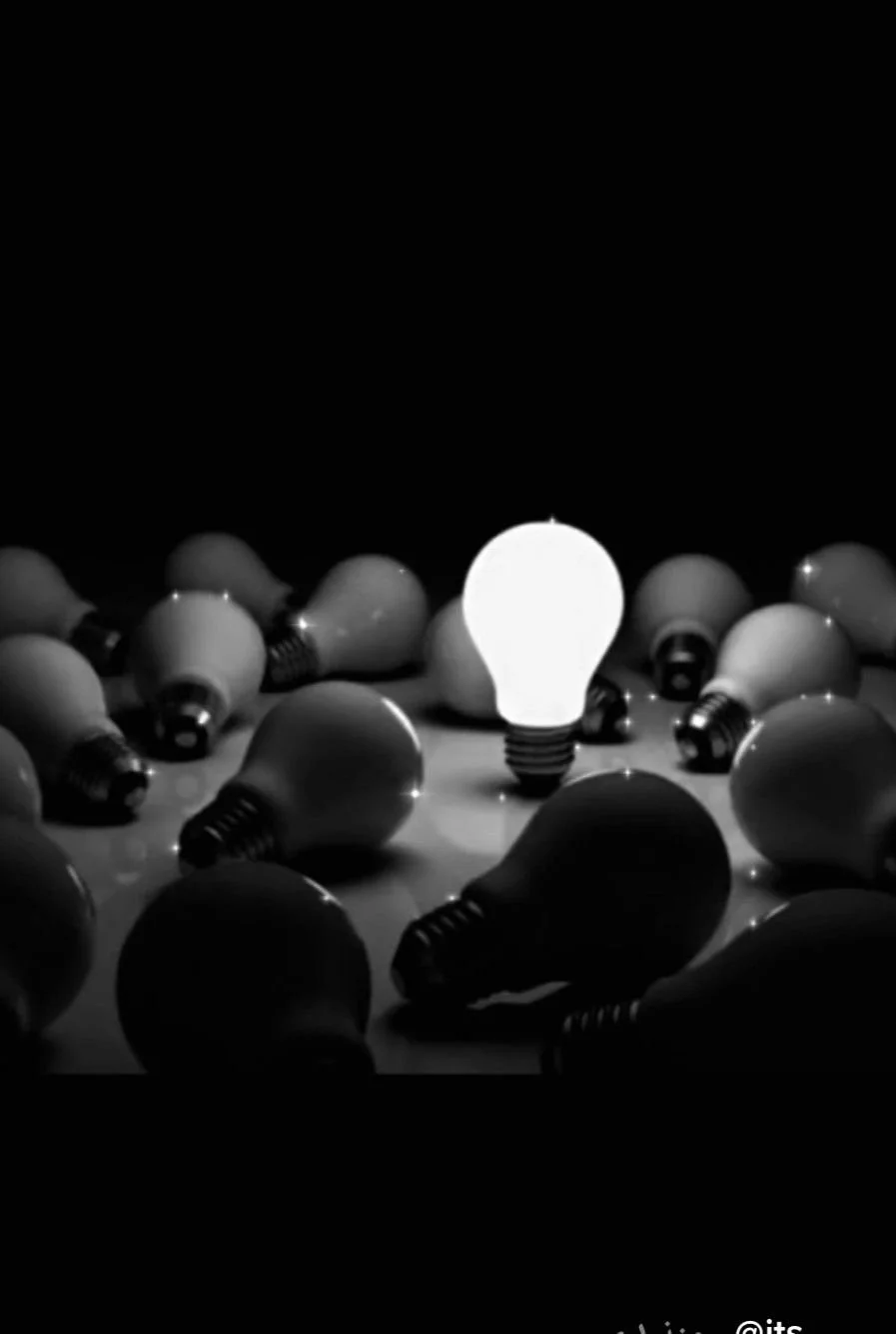 A lit light bulb among unlit light bulbs scattered on a surface, with a dark background.