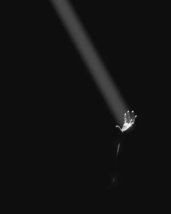 A black-and-white image of a hand reaching out with a bright beam of light shining down on it against a dark background.