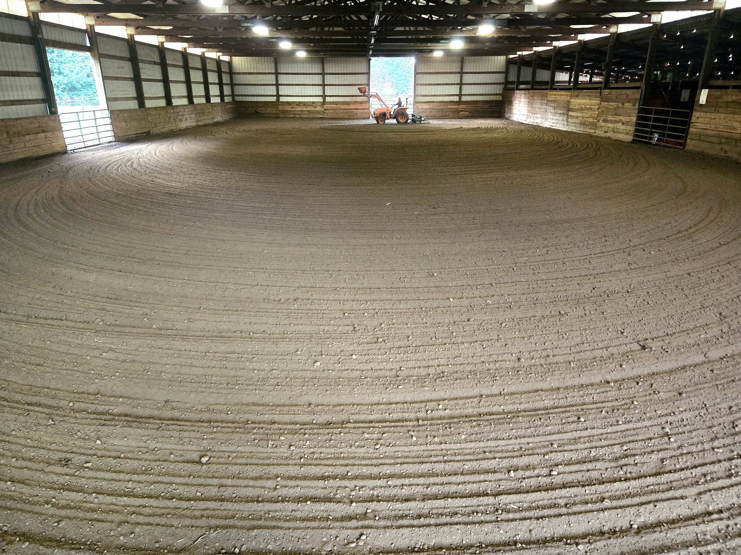 Indoor riding arena with a machine leveling dirt, wooden walls, some windows, and overhead lighting.