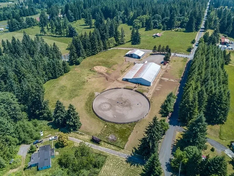 Aerial view of a rural property with stable buildings, a round riding arena, and surrounding green trees and fields.