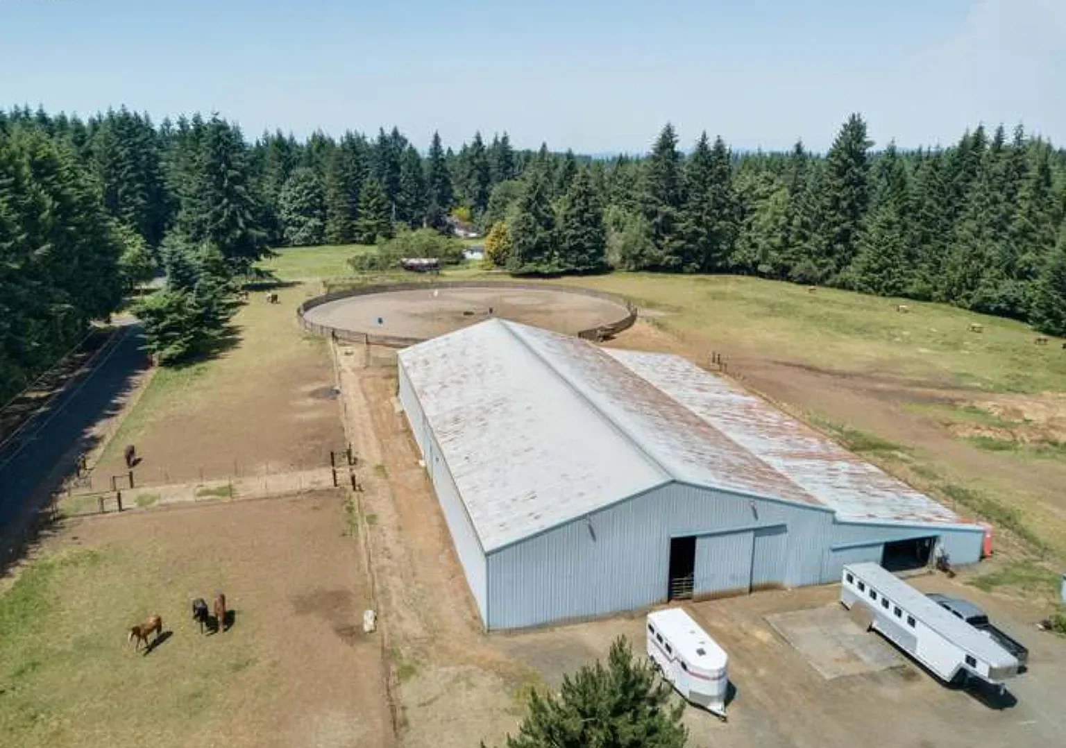 Aerial view of a large farm with a long metal barn, a horseshoe-shaped riding arena, fenced paddocks, and a camper trailer parked nearby, surrounded by trees and open fields.