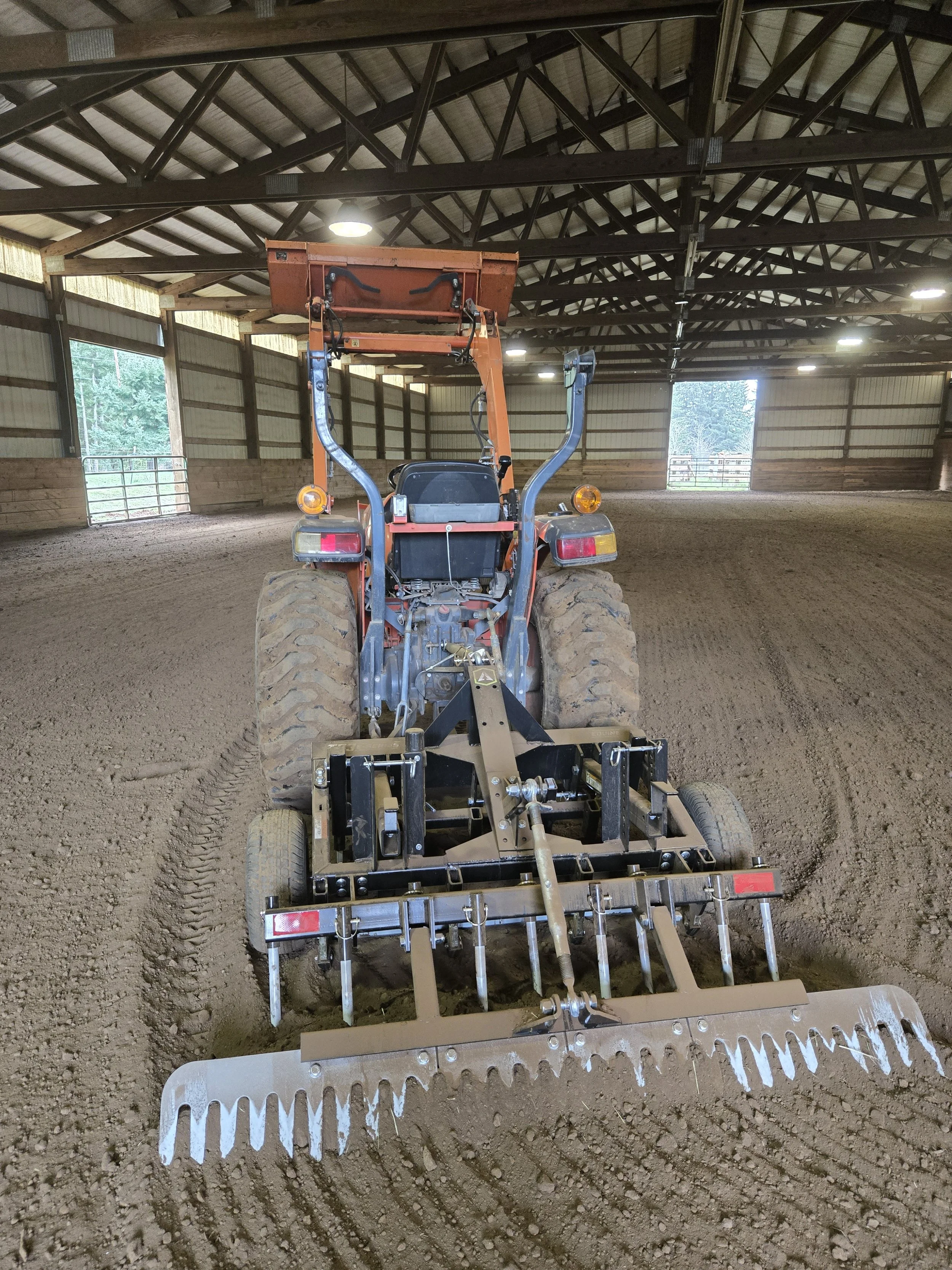 A tractor with a landscaping attachment grooming the dirt floor inside an indoor riding arena.