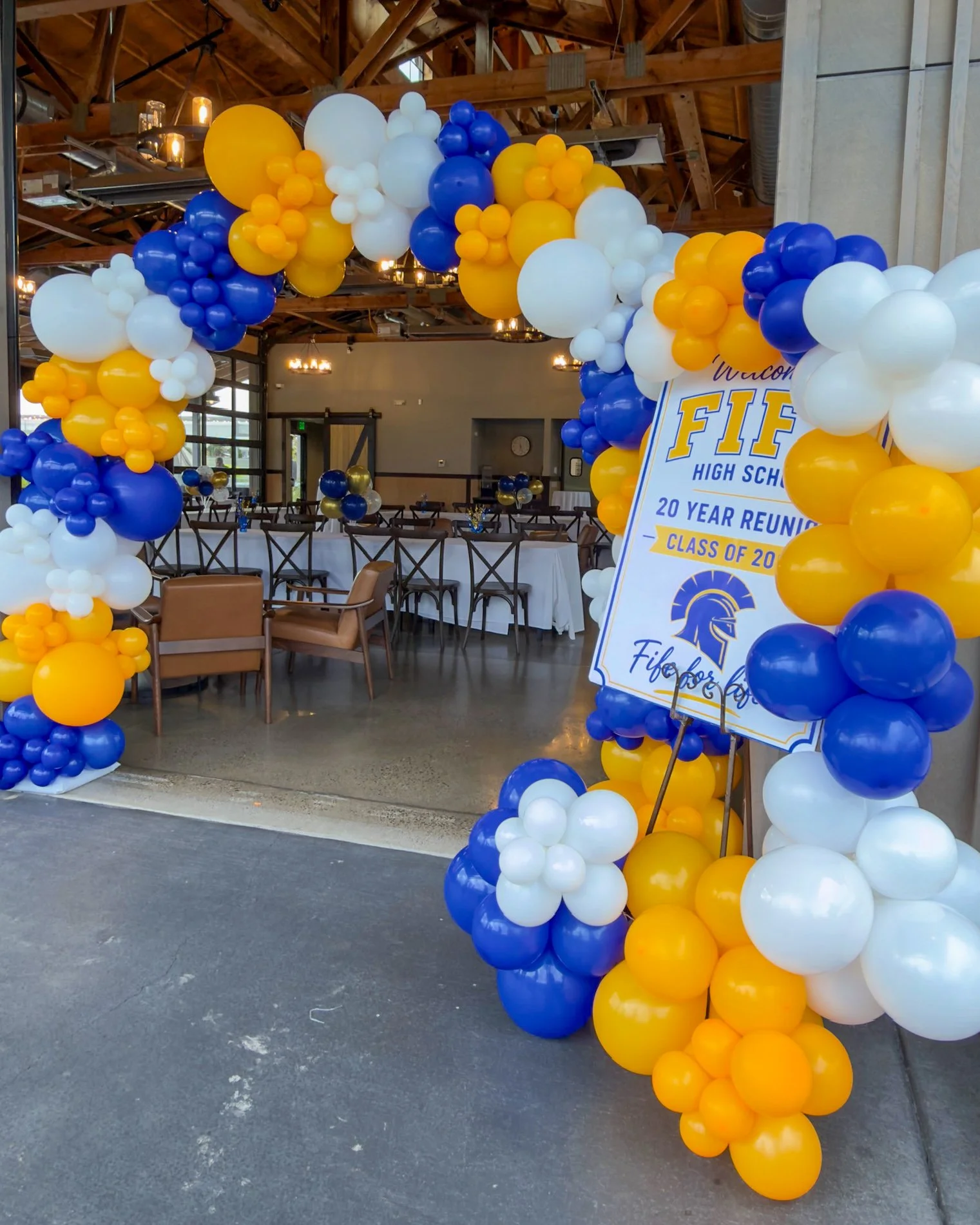 Decorative archway made of yellow, blue, and white balloons at a high school reunion event. Inside, tables are set up with chairs for guests. A sign indicates the celebration is for the Fife High School Class of 2003, marking a 20-year reunion.