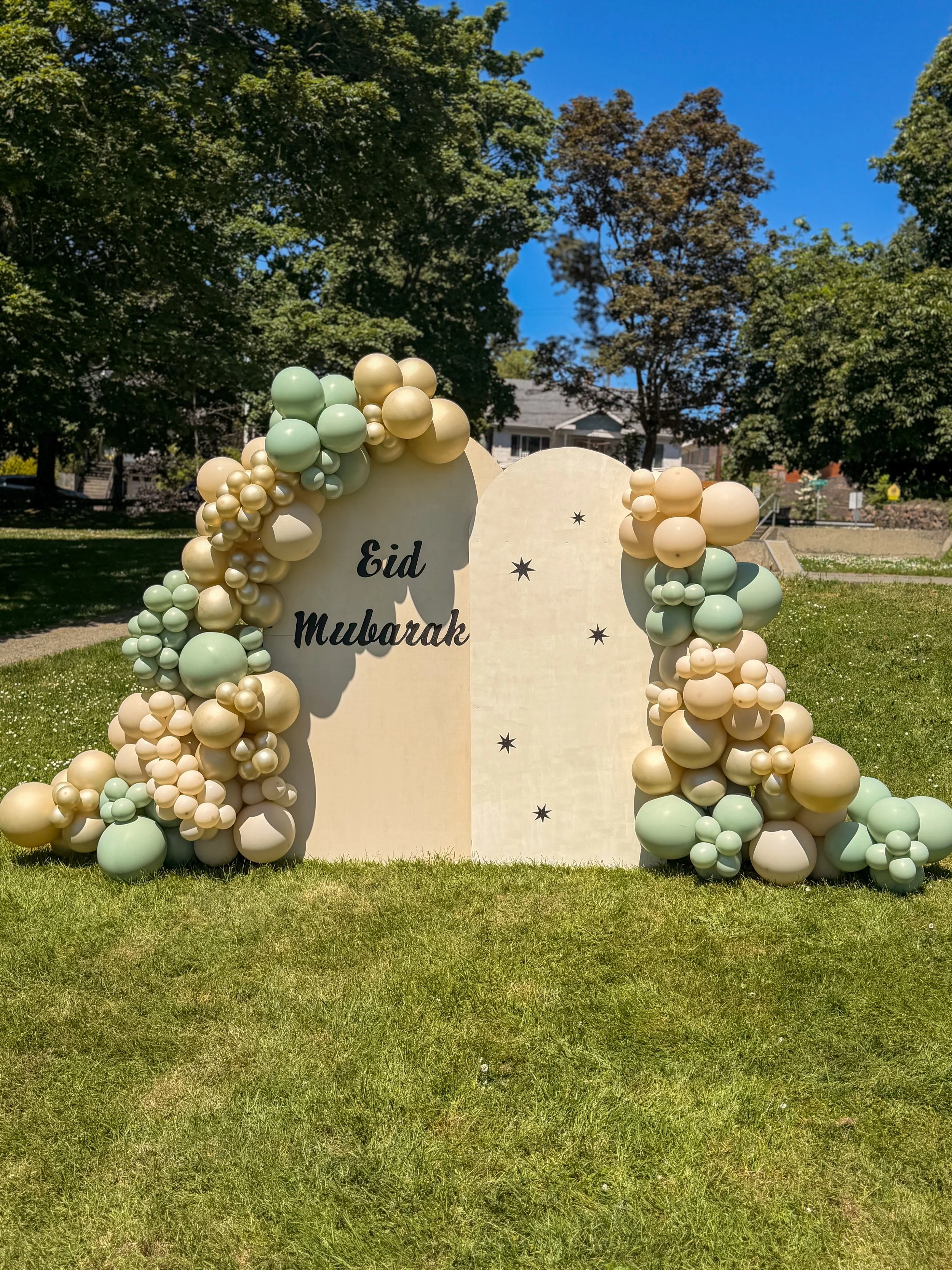 Decorative arch with cream and light green balloons and stars, with the words 'Eid Mubarak' in the center, set outdoors on green grass with trees and houses in the background.