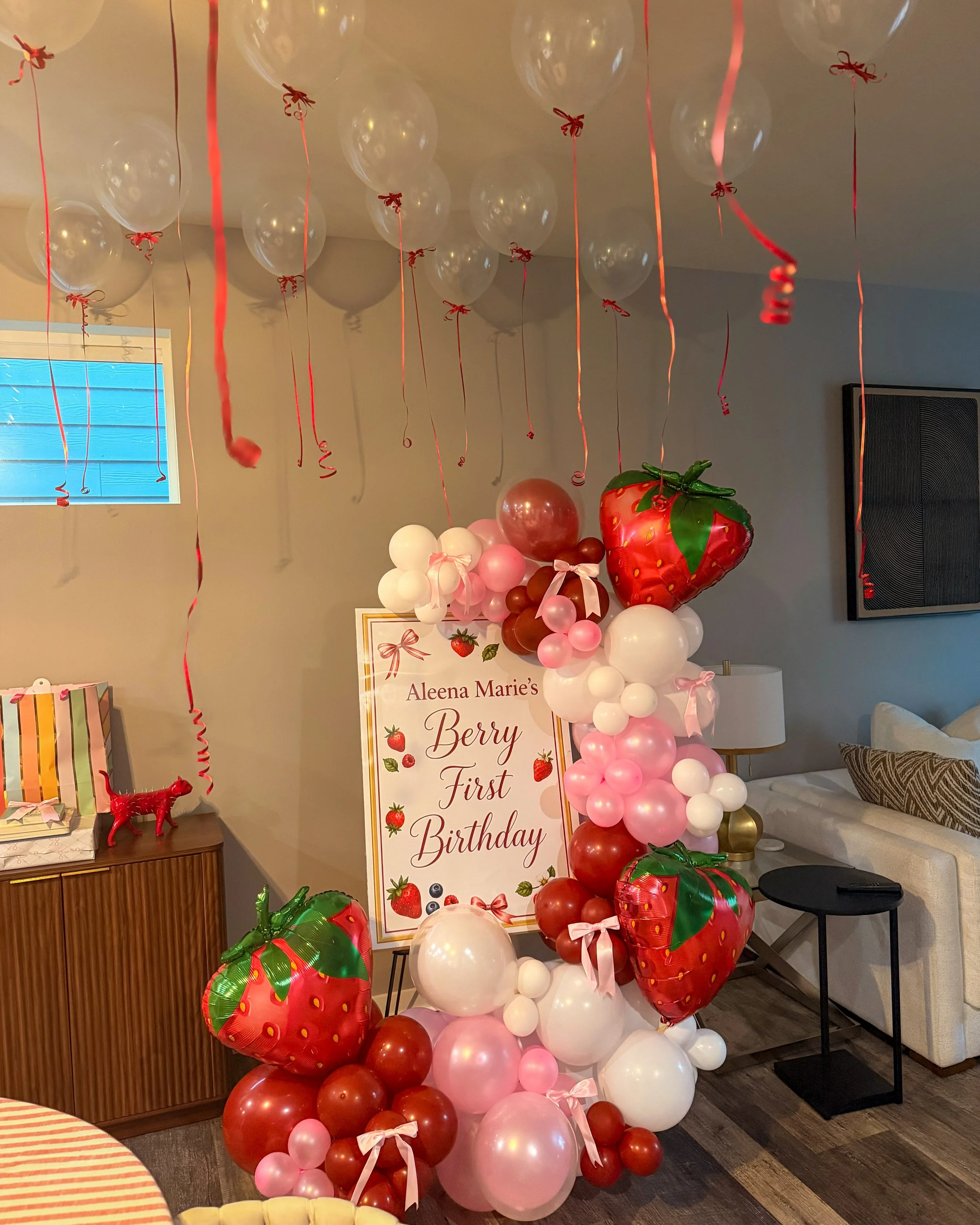 Decorative arrangement of balloons with strawberry theme for a first birthday celebration, includes a large strawberry-shaped balloon, a banner with strawberries and message, and pink, red, white balloons, with red ribbons hanging from the ceiling.