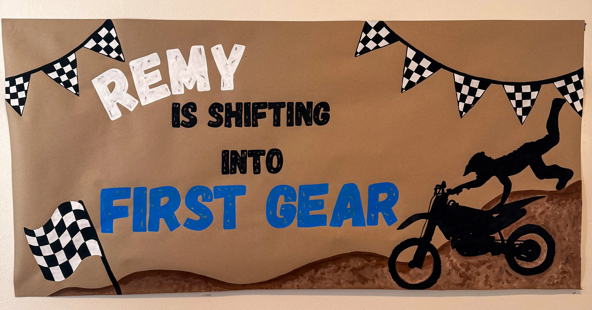 Decorative sign with racing flags and a silhouette of a person riding a dirt bike, saying 'Remy is shifting into first gear'.