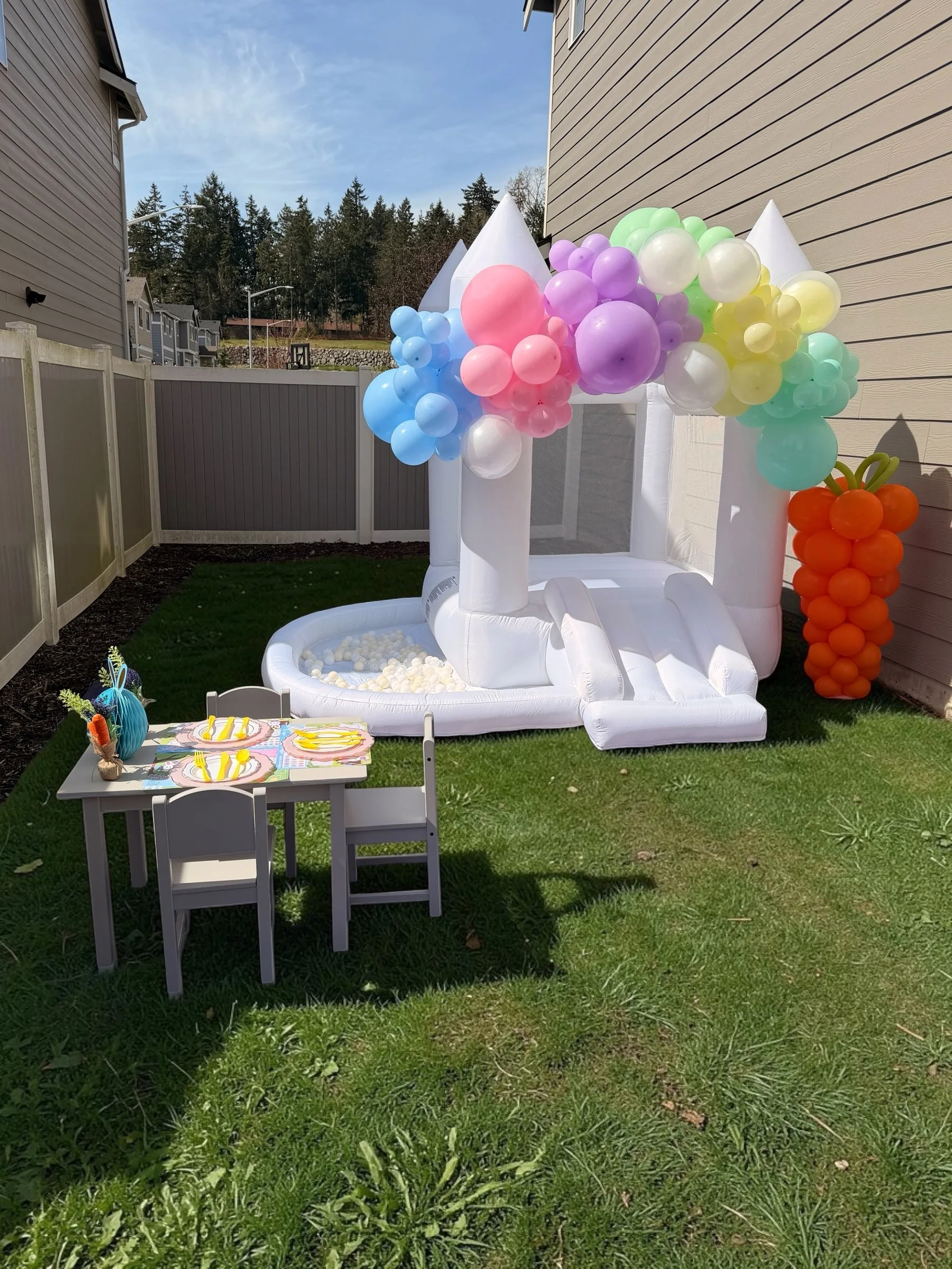 Inflatable baby birthday party setup with colorful balloons, a small table with plates and utensils, and a balloon carrot on the grass in a backyard.