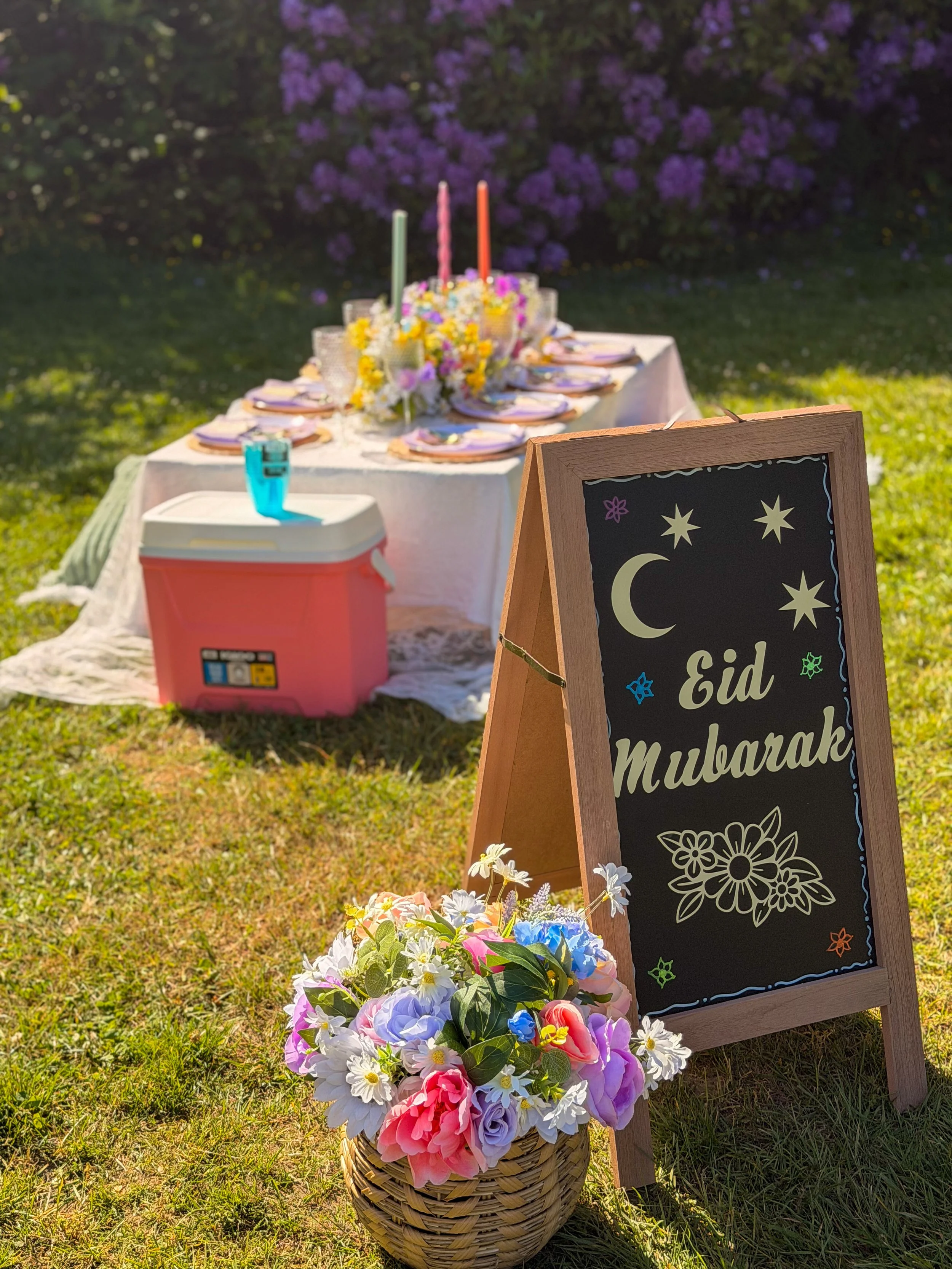 Easter celebration setup with a basket of colorful flowers, a decorated chalkboard sign reading 'Eid Mubarak', and a table with plates and candles in an outdoor garden setting.