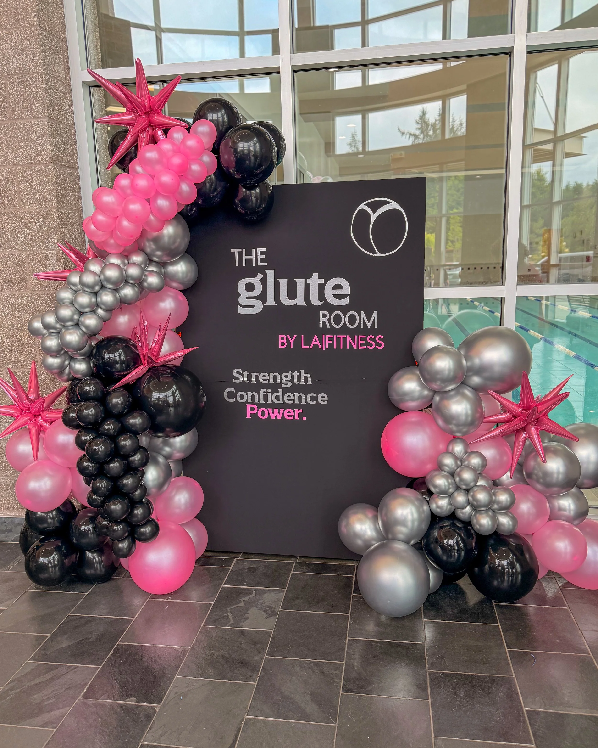 Decorative balloon arrangement with pink, black, and silver balloons and pink starburst-shaped balloons in front of a black sign with white and pink text for The Glute Room by LA Fitness at an indoor swimming pool.