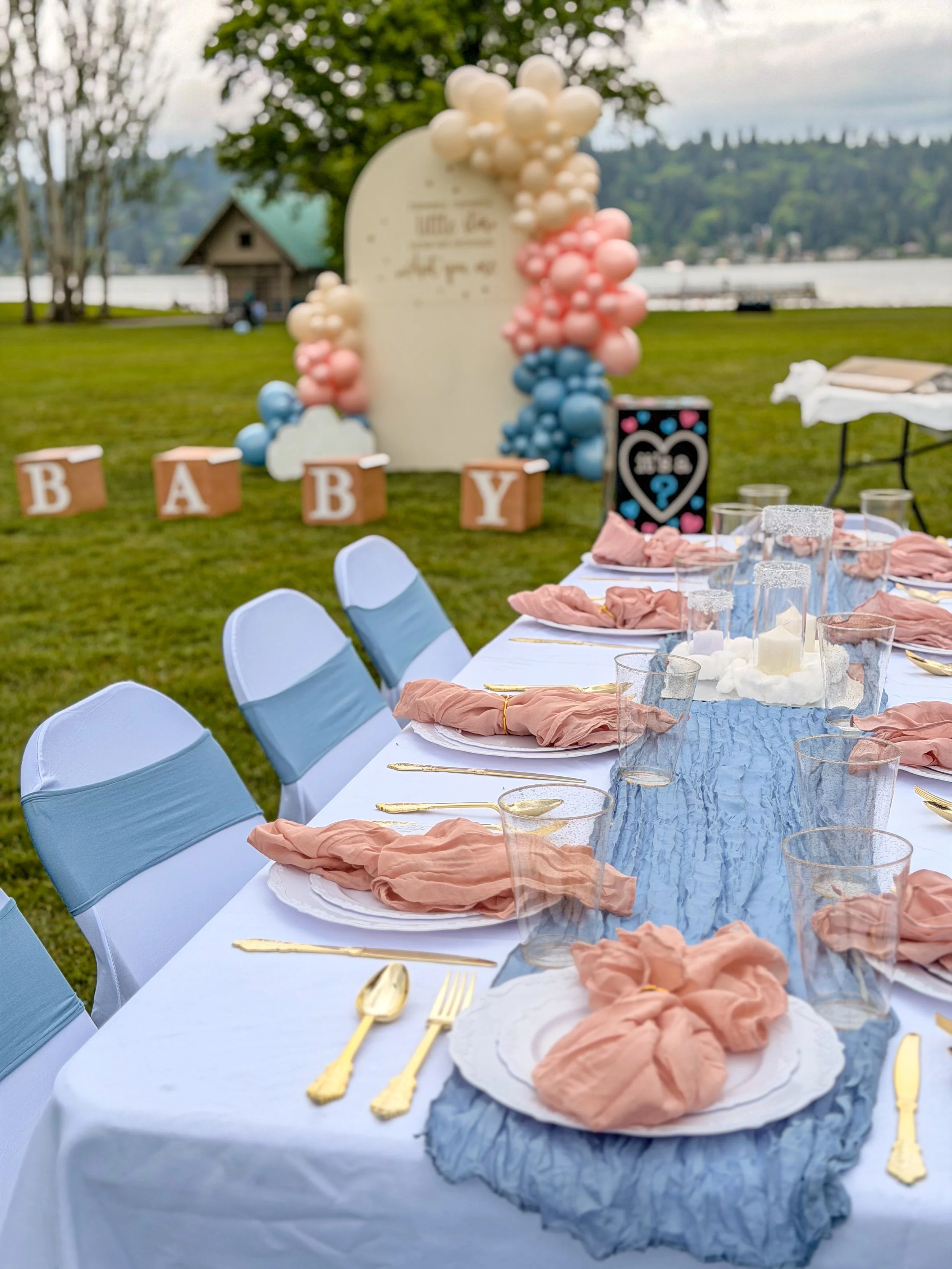 Long outdoor table set for a celebration near a lake with blank chairs, gold utensils, pink napkins, and a blue table runner, with a backdrop of trees, a small building, and a lake view.