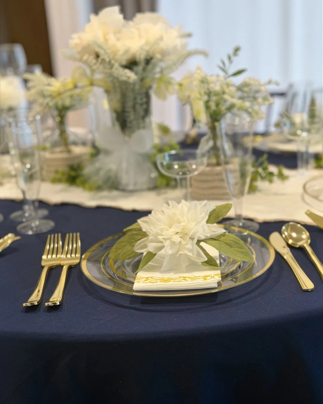 Elegant dining table with navy blue tablecloth, gold cutlery, a clear glass plate with a white flower and green leaves, and white floral centerpieces in vases with greenery.
