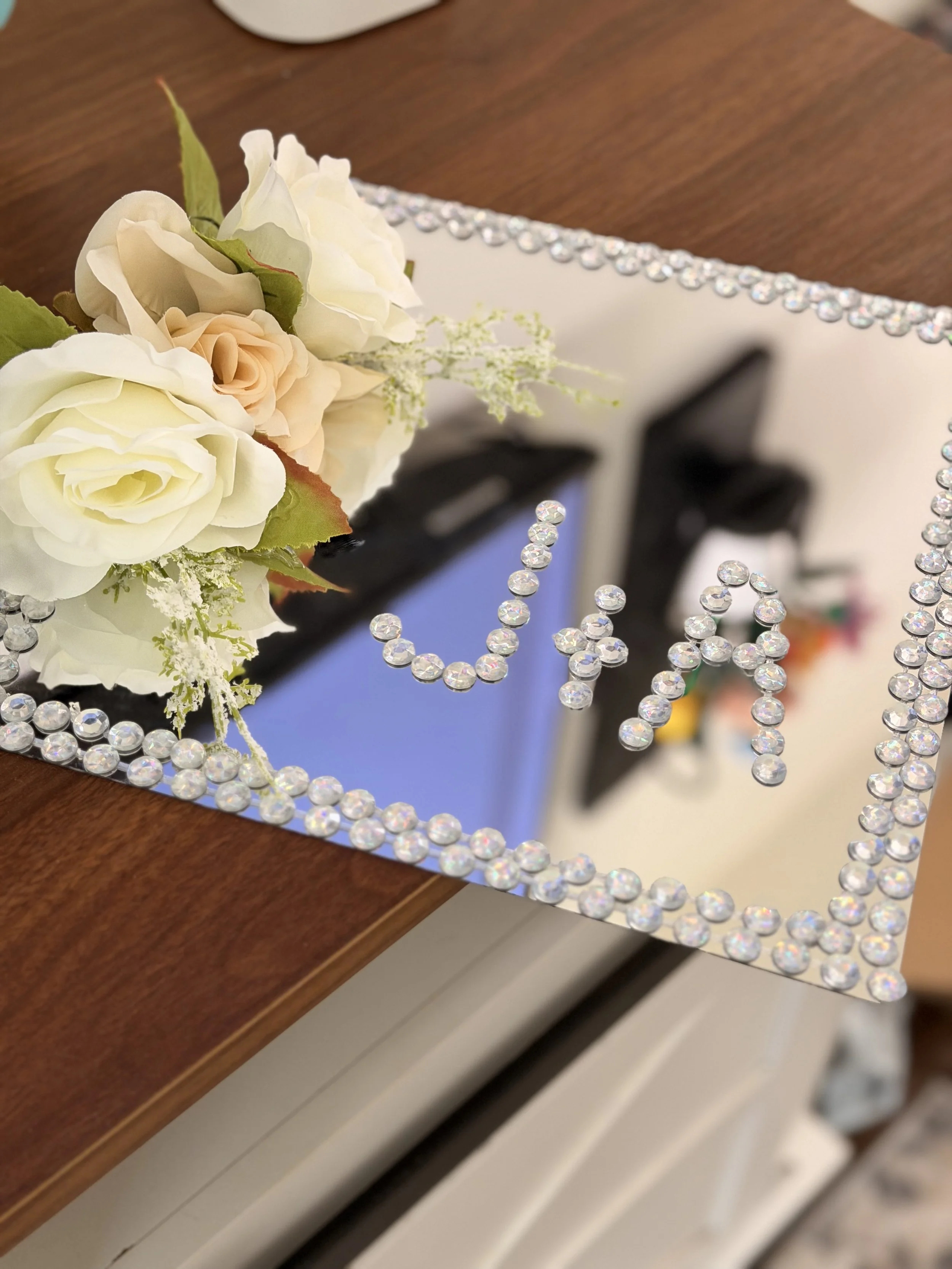 A bouquet of white and pale peach flowers on a wooden surface, with a decorative mirror with rhinestones around its edges, reflecting piano keys below.