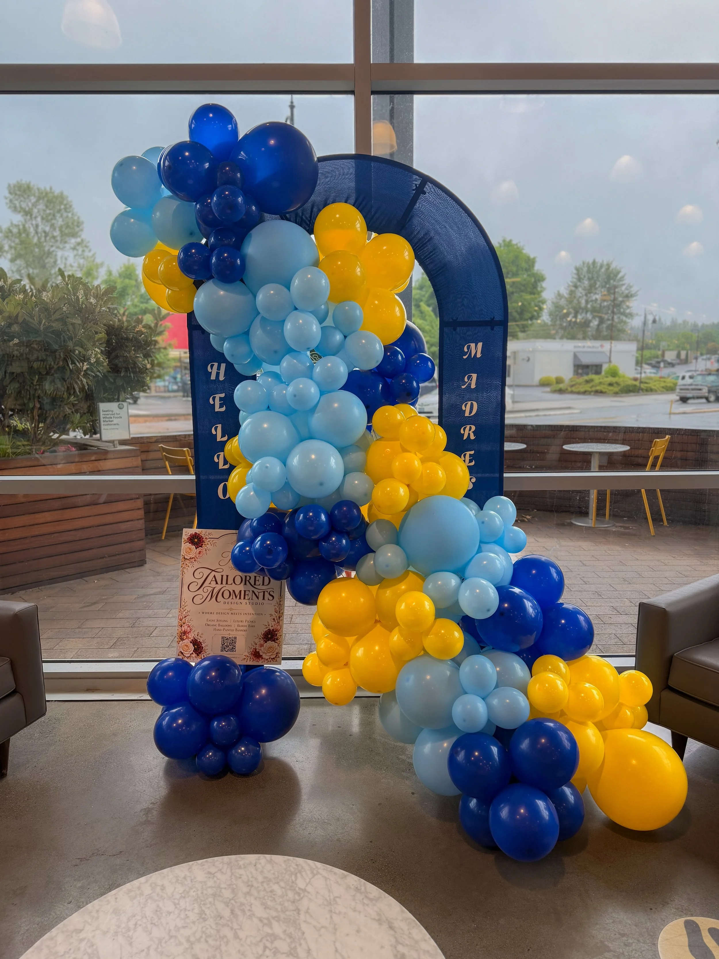 Decorative balloon arch with blue, light blue, and yellow balloons near a large window at an indoor event space.