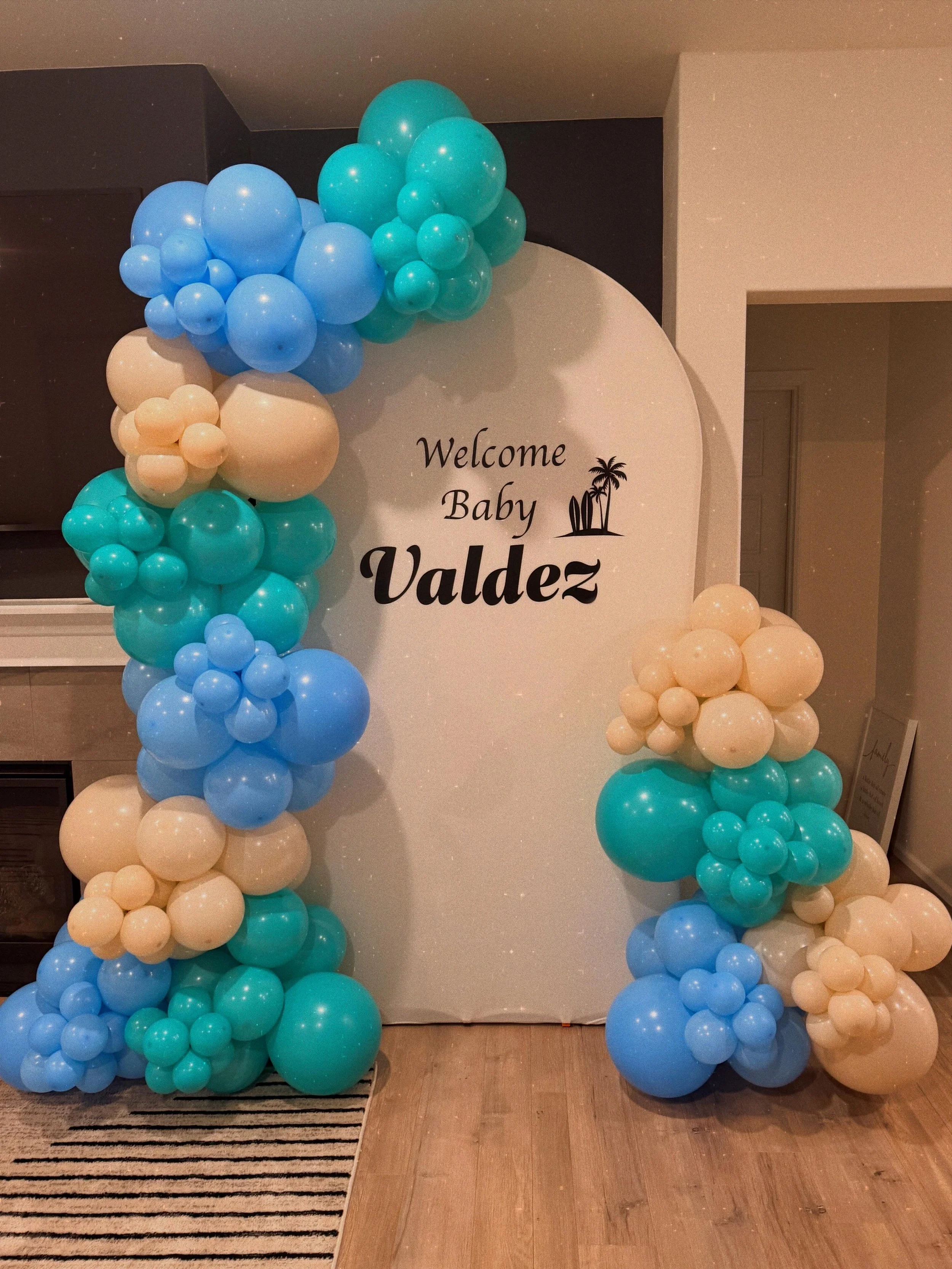 Decorative arch made of blue, teal, and beige balloons with a sign that says "Welcome Baby Valdez" and a small palm tree graphic.
