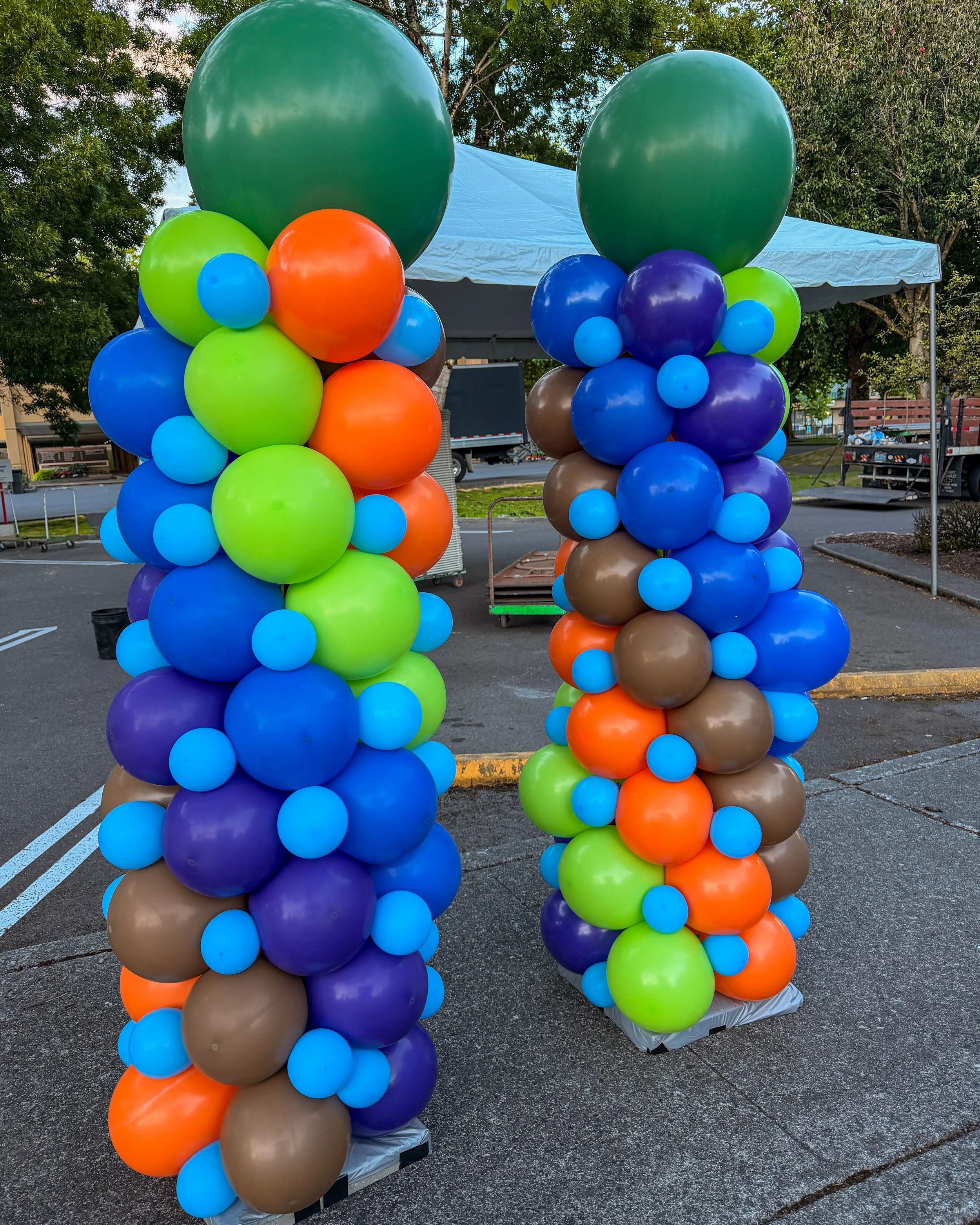 Colorful balloon columns with large green balloons on top, set up outdoors near a tent and trees.