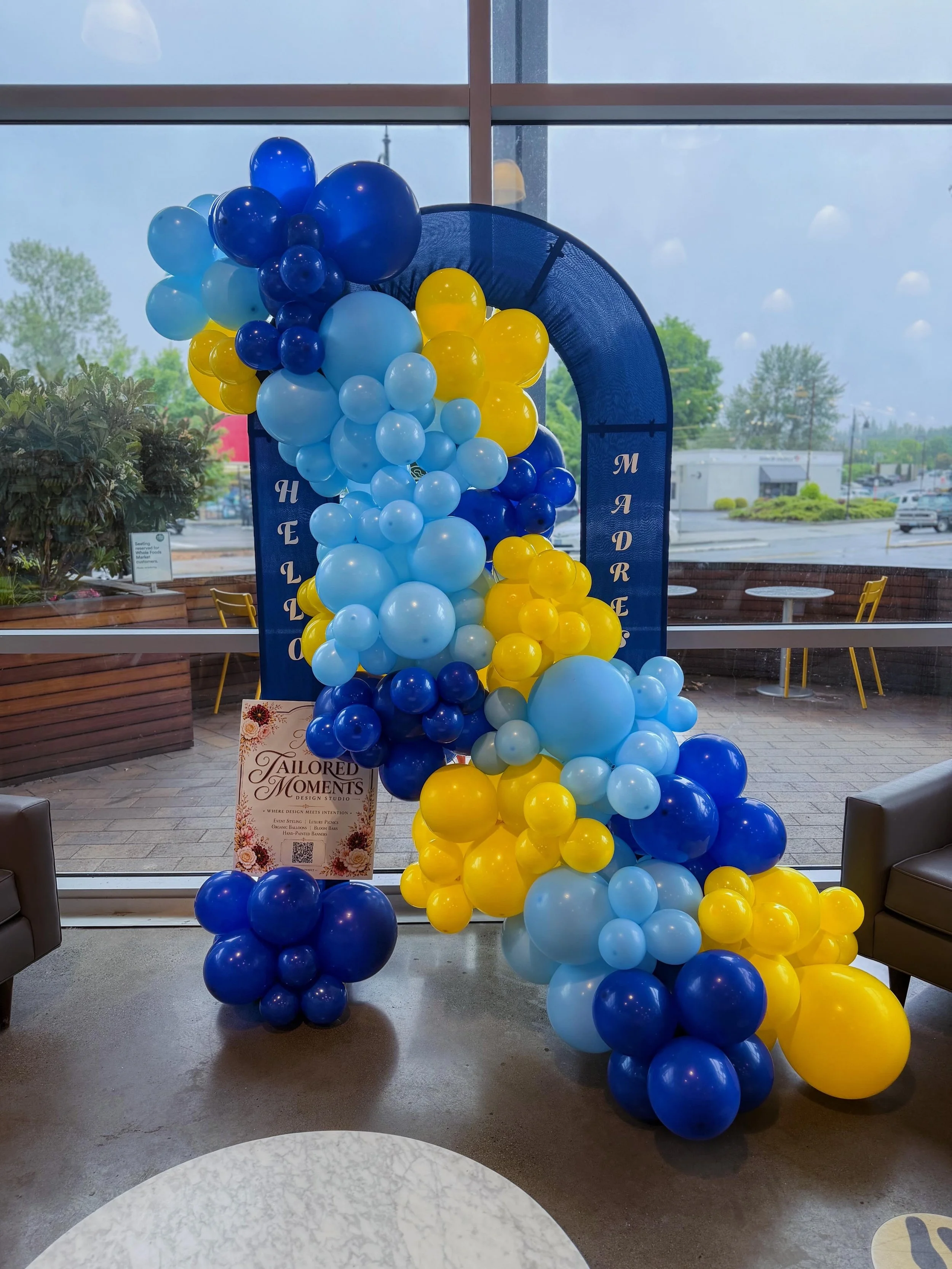 A large decorative balloon display with yellow, light blue, and dark blue balloons arranged in a cascading pattern in front of a window. The window shows an outdoor parking lot and trees. A sign nearby reads "Tailored Moments Design Studio."