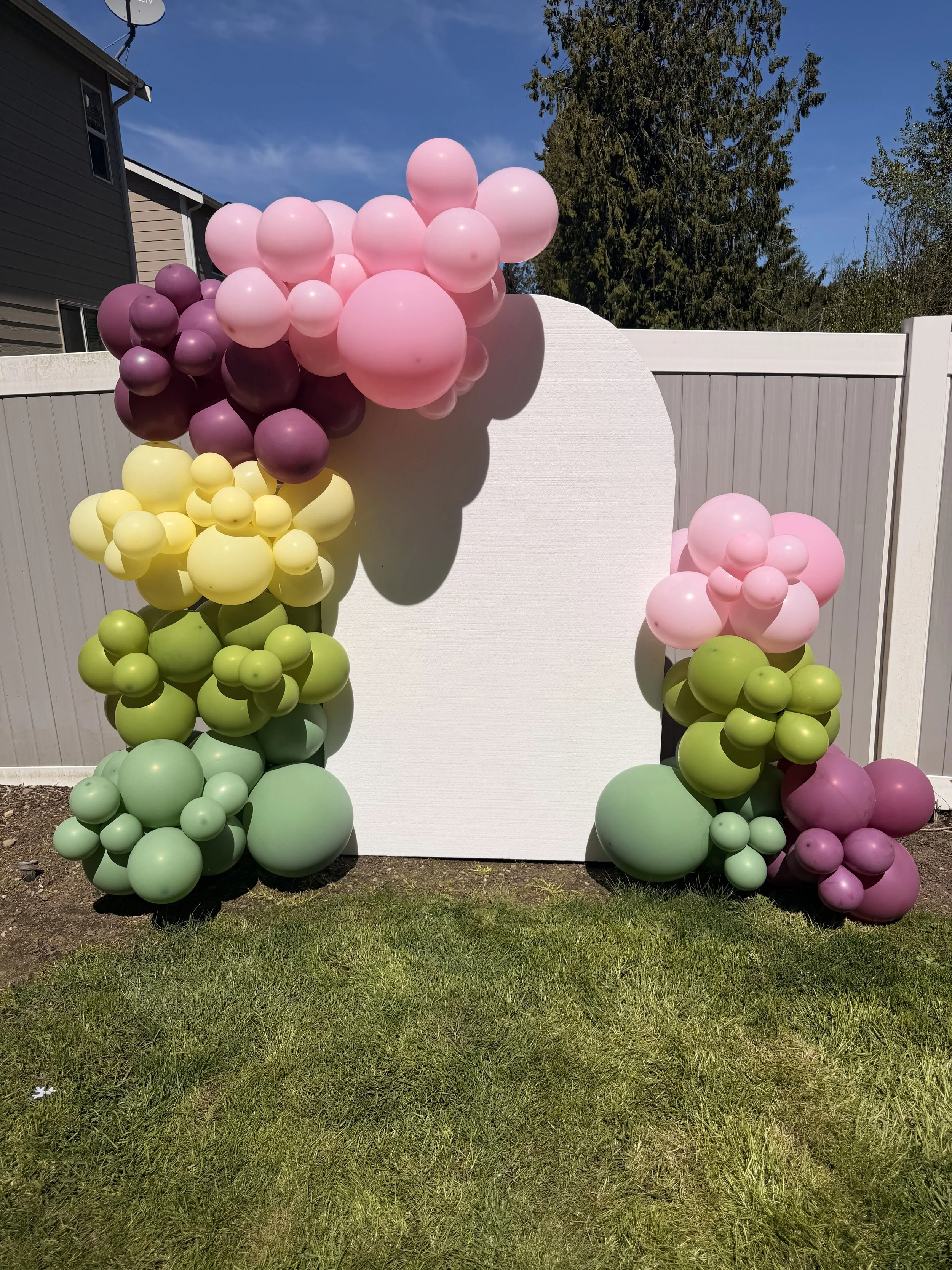 Colorful balloon decorations with pastel pink, purple, yellow, and green balloons arranged around a white backdrop outdoors on a sunny day.