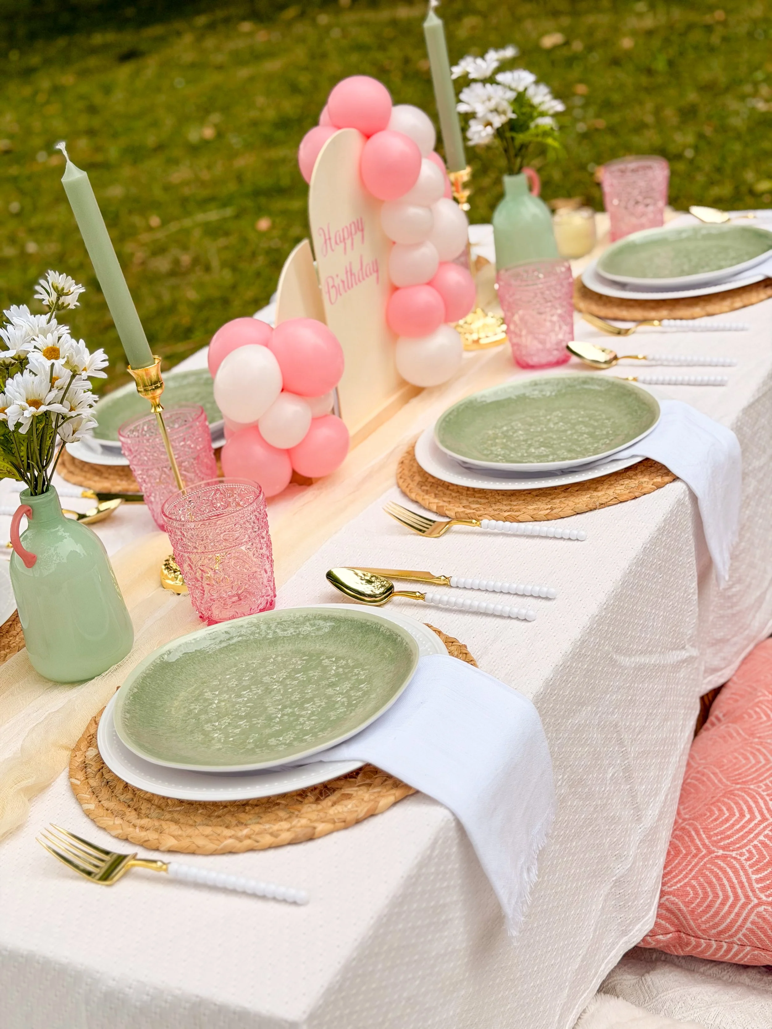 A table set for a celebration with pink and green themed decorations, balloons, candles, flowers, plates with green soup, and gold utensils.