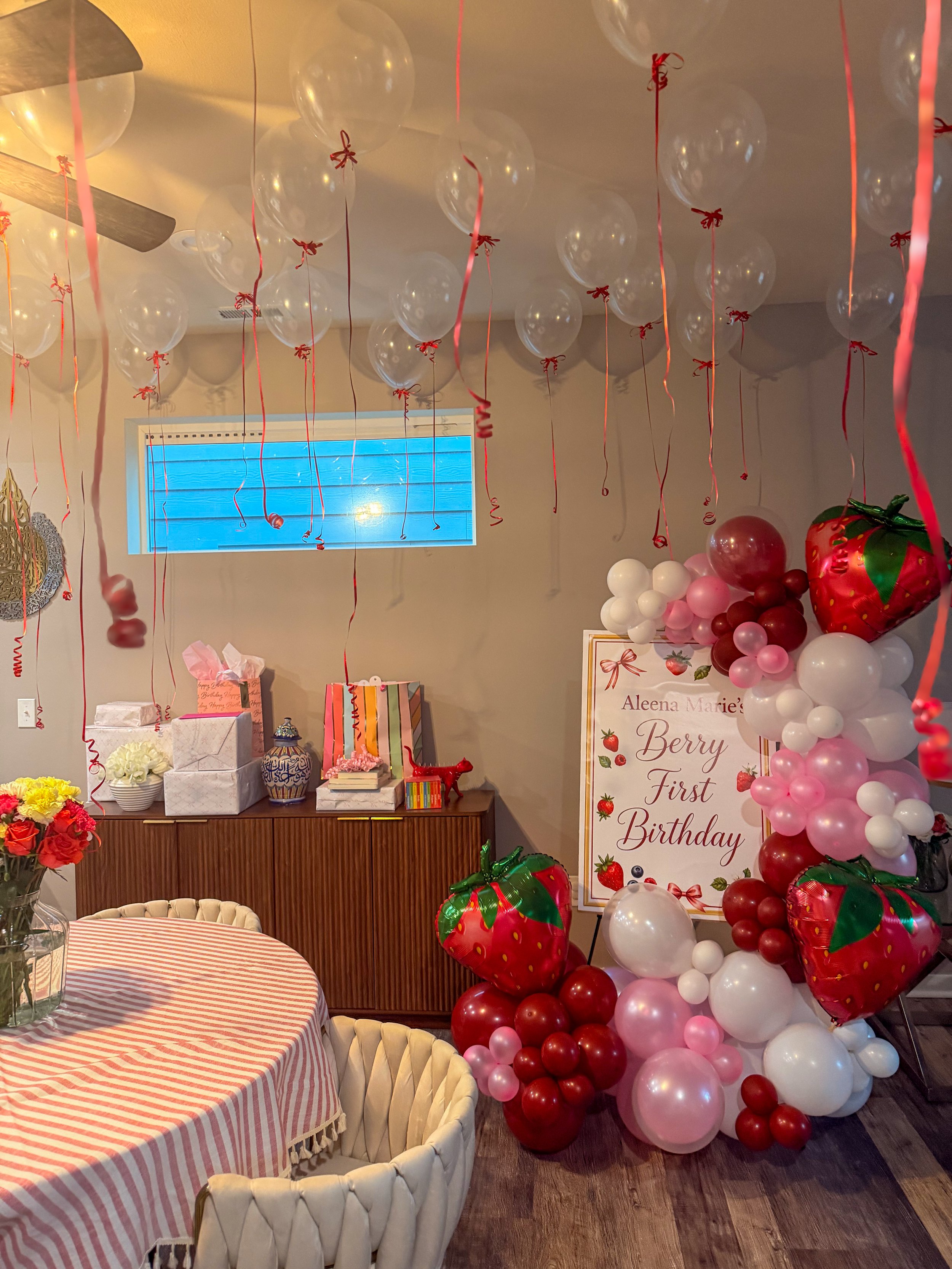Decorations for a birthday party including pink, red, and white balloons arranged in clusters, a large strawberry-shaped balloon, a birthday sign that reads 'Aleena Marie's Berry First Birthday,' a table with a pink striped tablecloth and a flower arrangement, gift boxes on a sideboard, and a wall with a small window and wall decorations.