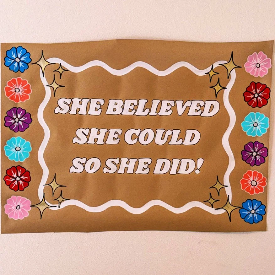 A motivational poster with a brown background, decorated with colorful flowers and stars, that says 'She believed she could so she did!' in white letters.