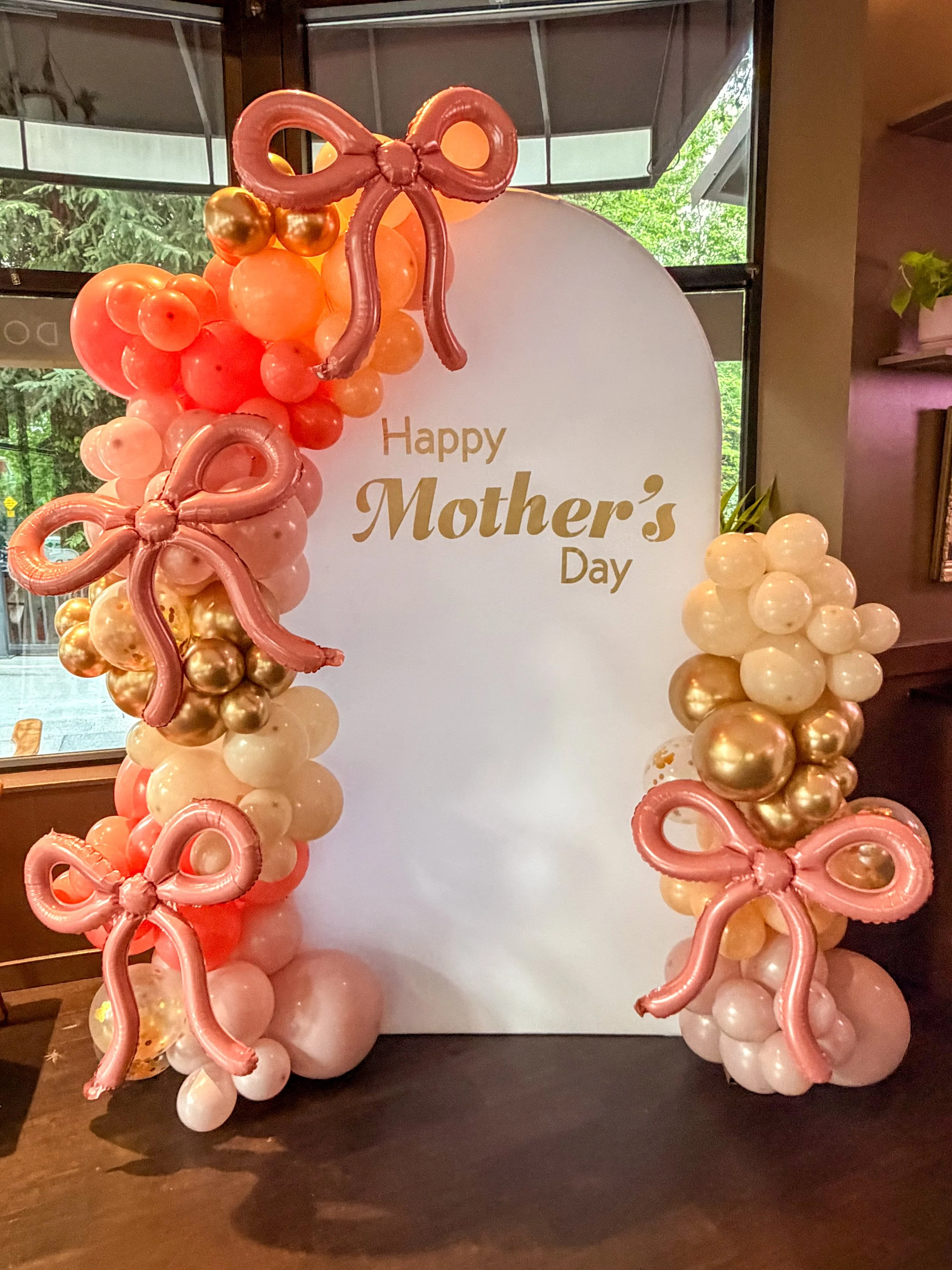 Balloon arch with pink, gold, and white balloons and pink balloon butterflies, with a White backdrop that says 'Happy Mother's Day' in gold text, set up indoors near a window with greenery outside.