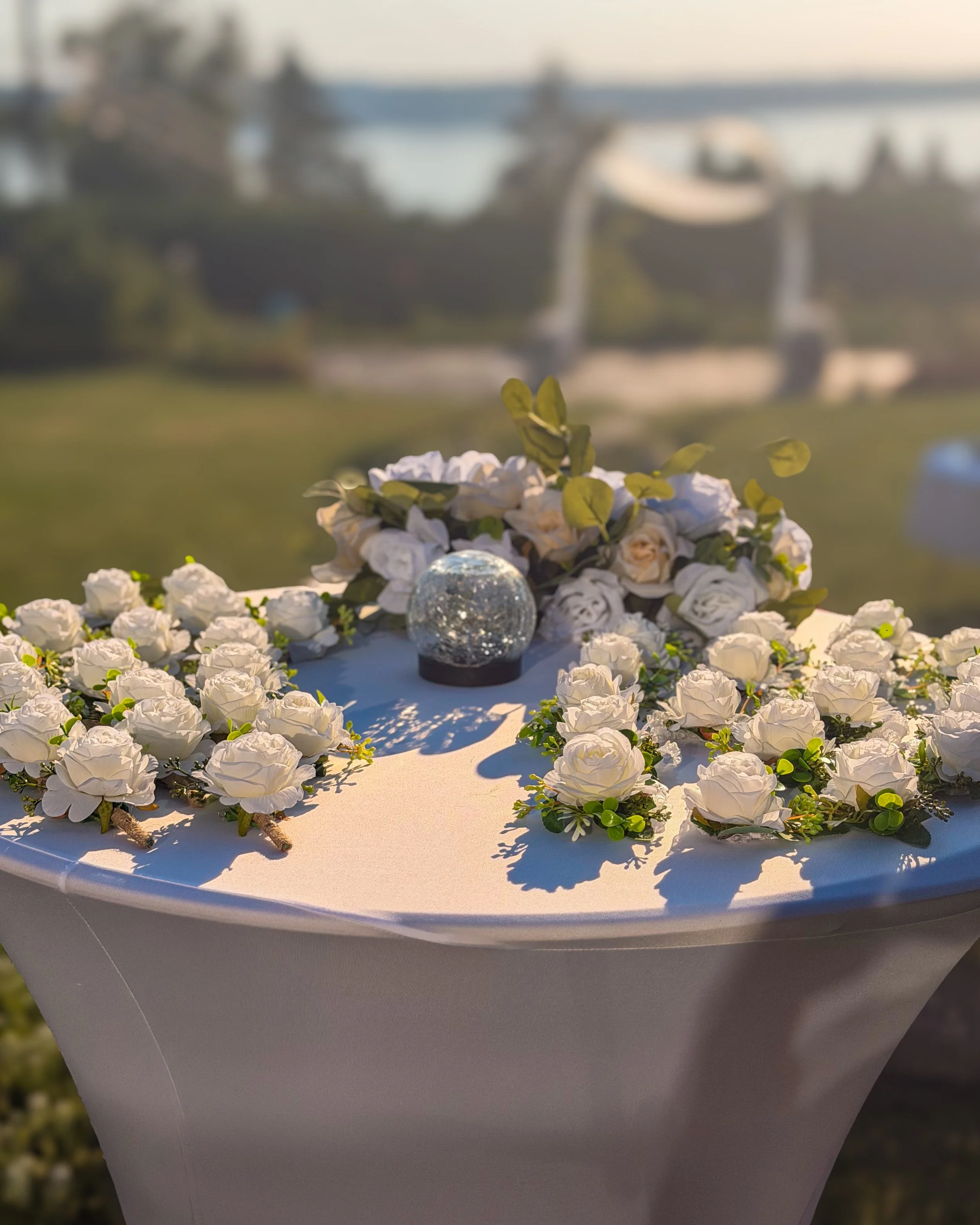 Close-up of a floral arrangement with white roses and greenery on a round table, with a blurred background of a bridge and trees.