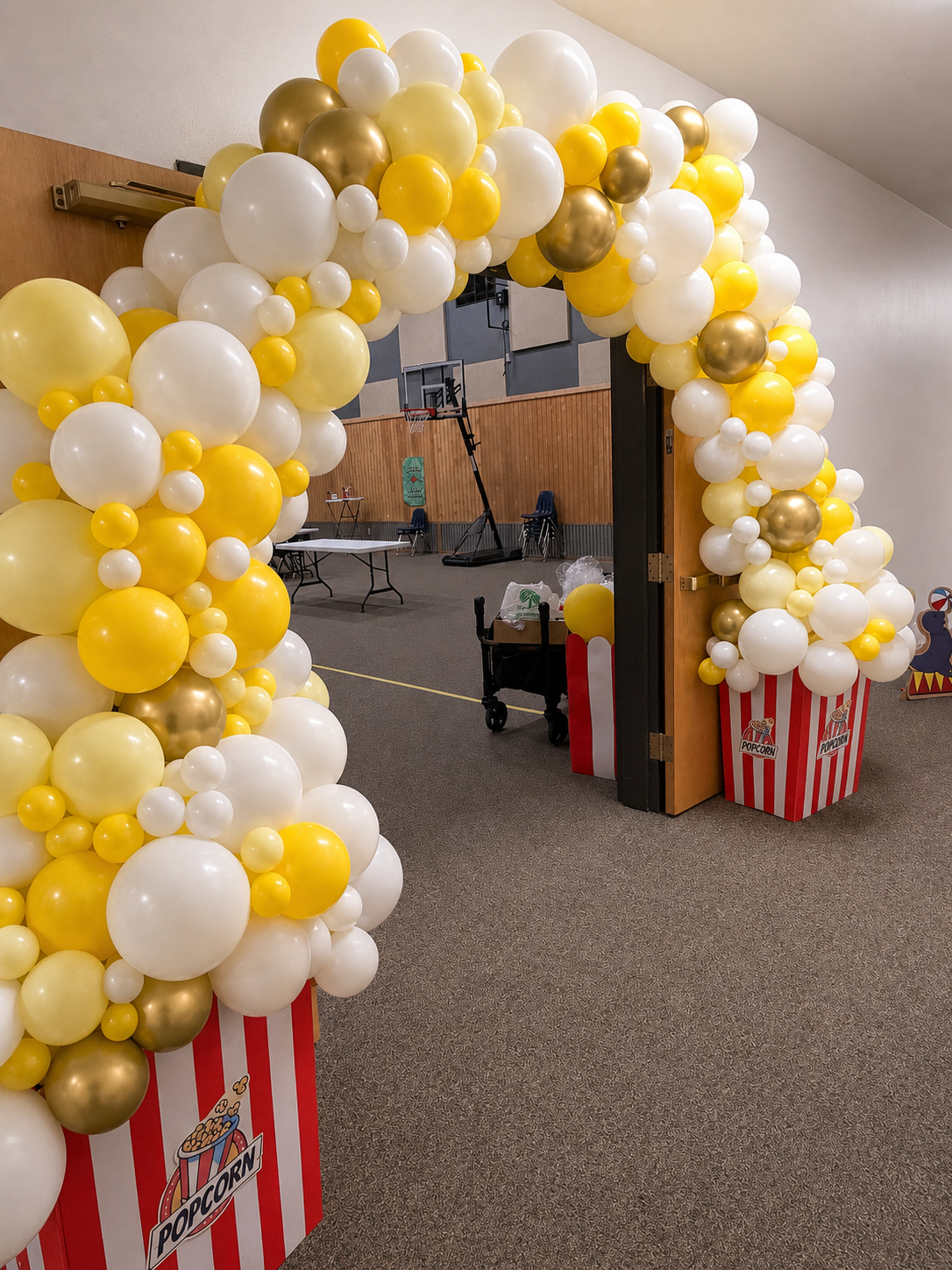 Balloon arch in yellow, white, and gold around an open doorway decorated with popcorn boxes. Inside, a gymnasium with basketball hoops, chairs, and tables is visible.