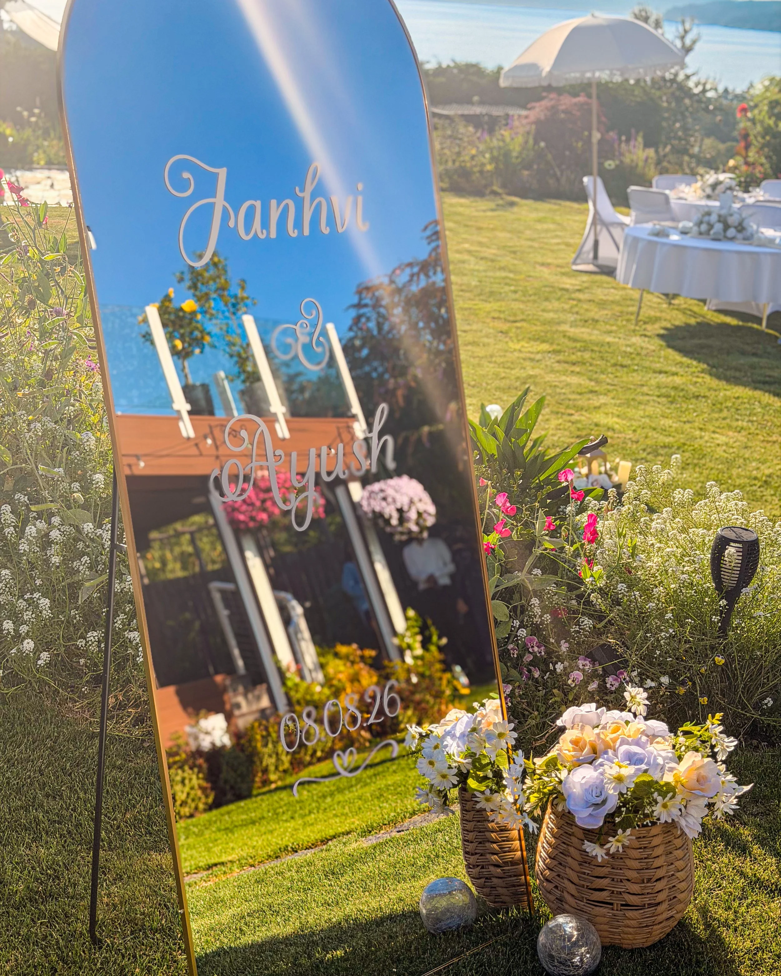 Outdoor wedding setup with a sign displaying the names "Janhvi" and "Aryush," surrounded by flower arrangements in baskets and decorative glass balls on the grass, with tables and an umbrella in the background under clear skies.
